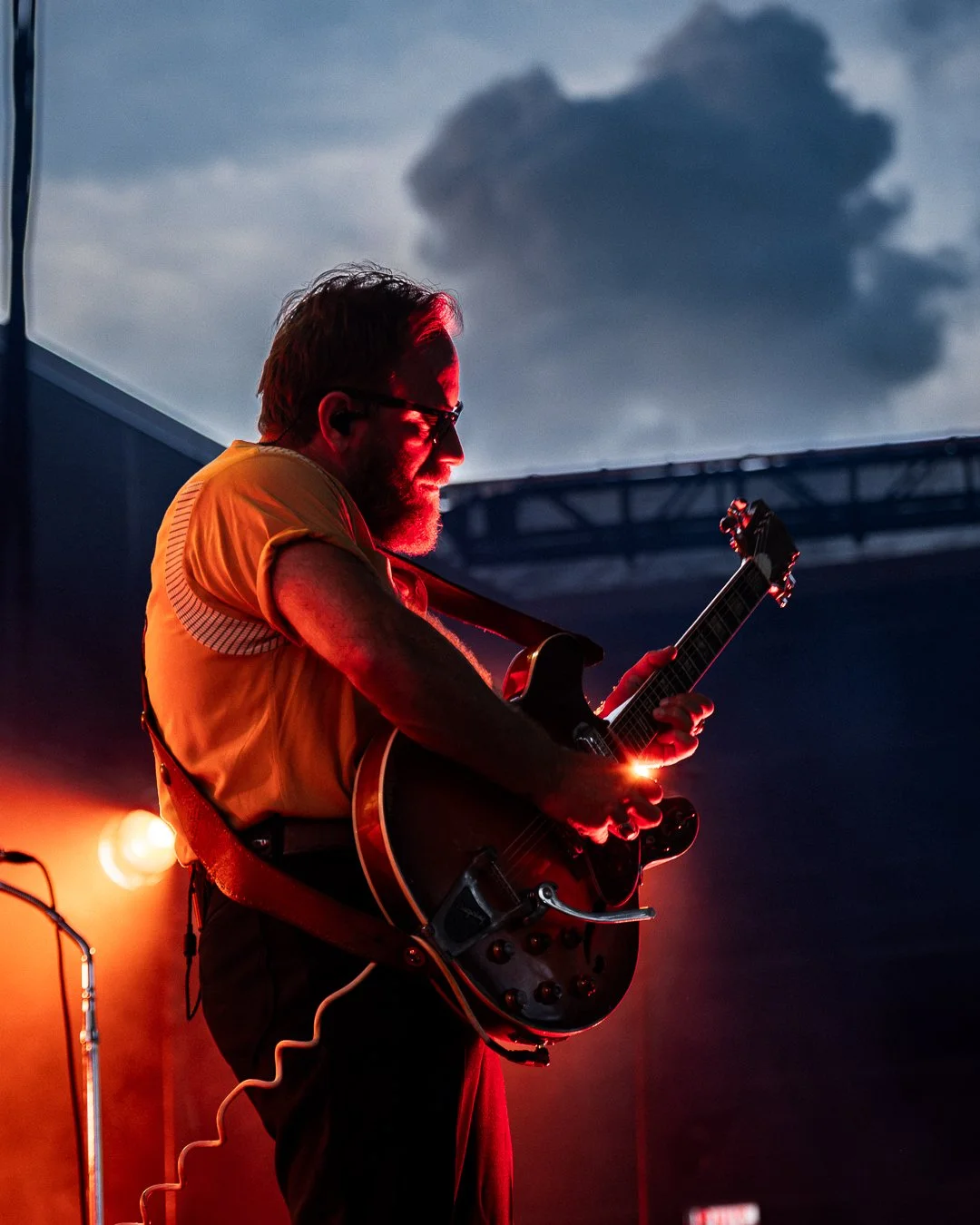 A male musician playing an electric guitar on stage during a live performance, with dark clouds visible in the evening sky behind him.