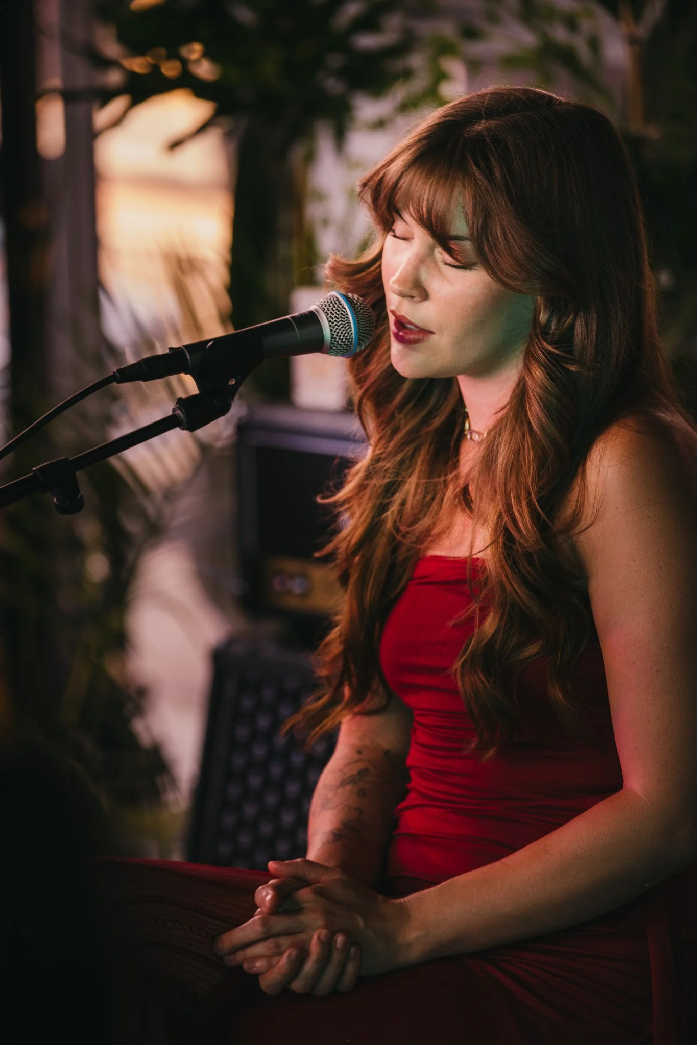 A woman with long wavy brown hair, wearing a red dress, is singing into a microphone with her eyes closed and hands clasped, in a warm dimly lit setting.