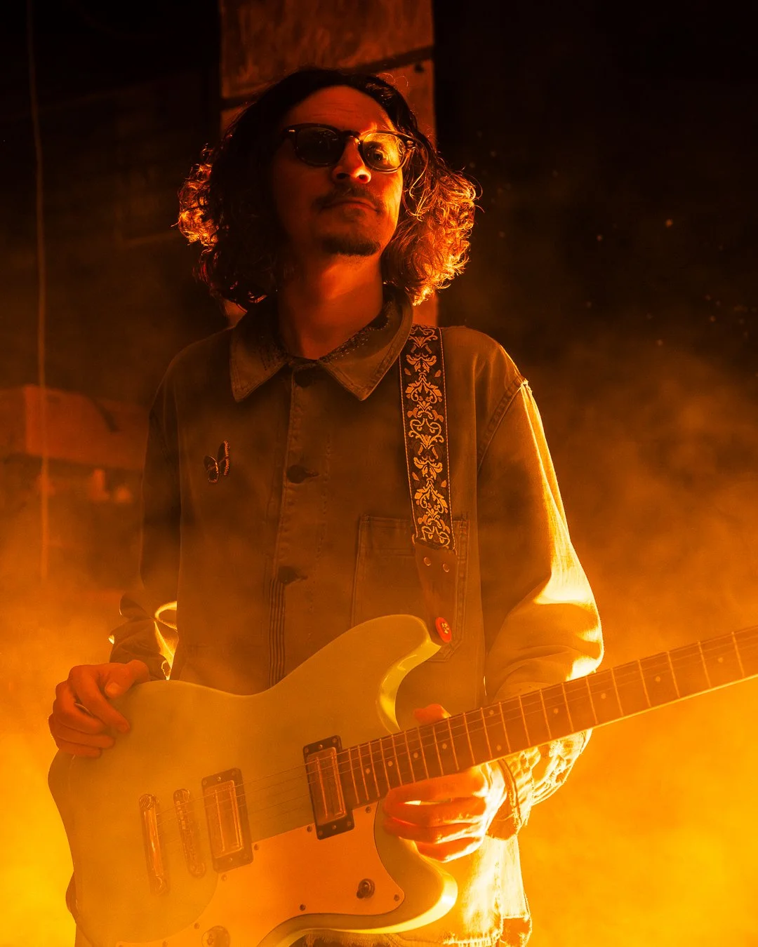 A musician with shoulder-length curly hair, wearing sunglasses and a denim jacket, playing a white electric guitar on stage illuminated by warm orange lighting.