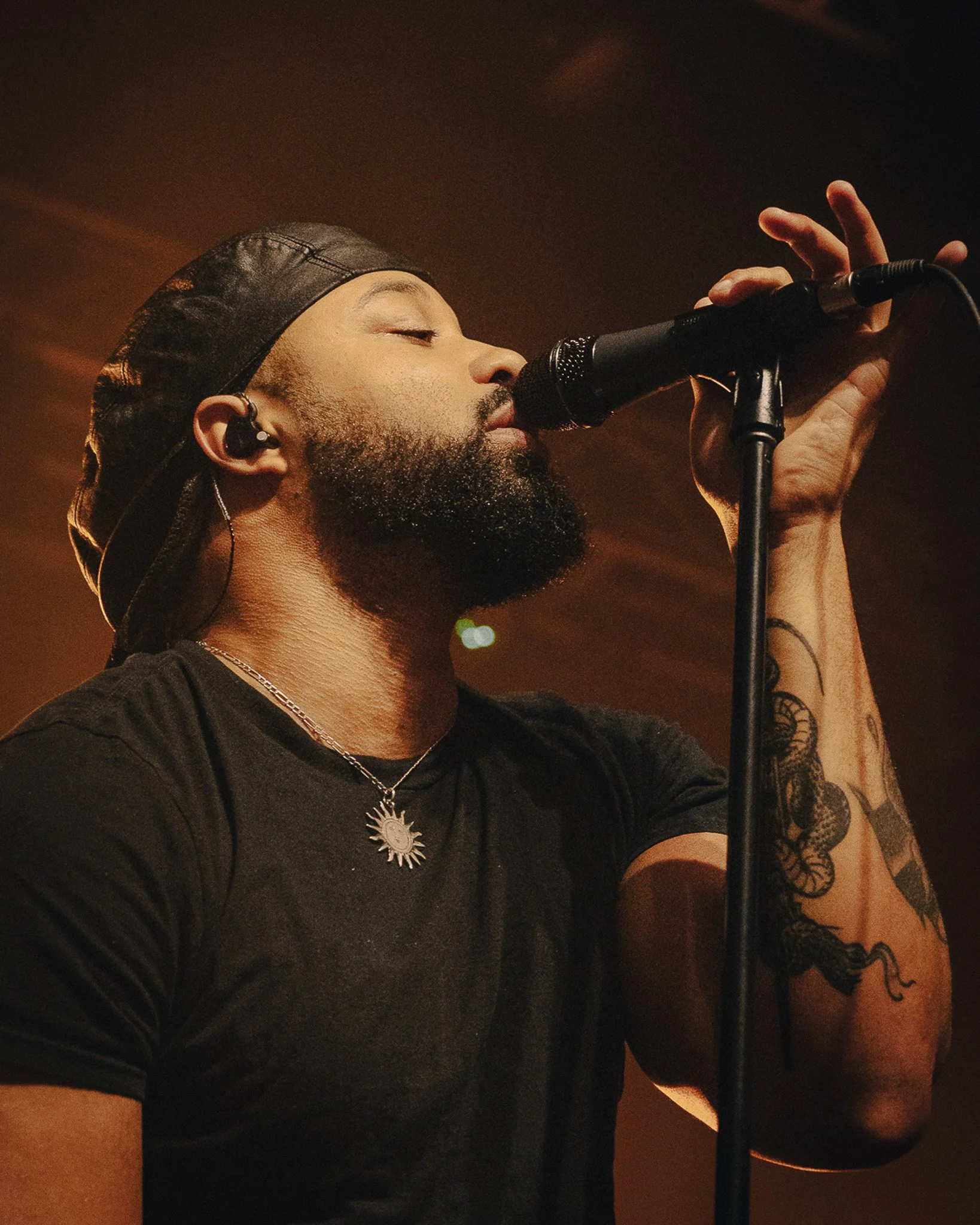 A male singer with a beard, wearing a leather cap, black shirt, a silver necklace, and earrings, is singing into a microphone with his eyes closed and holding the microphone stand with one hand.