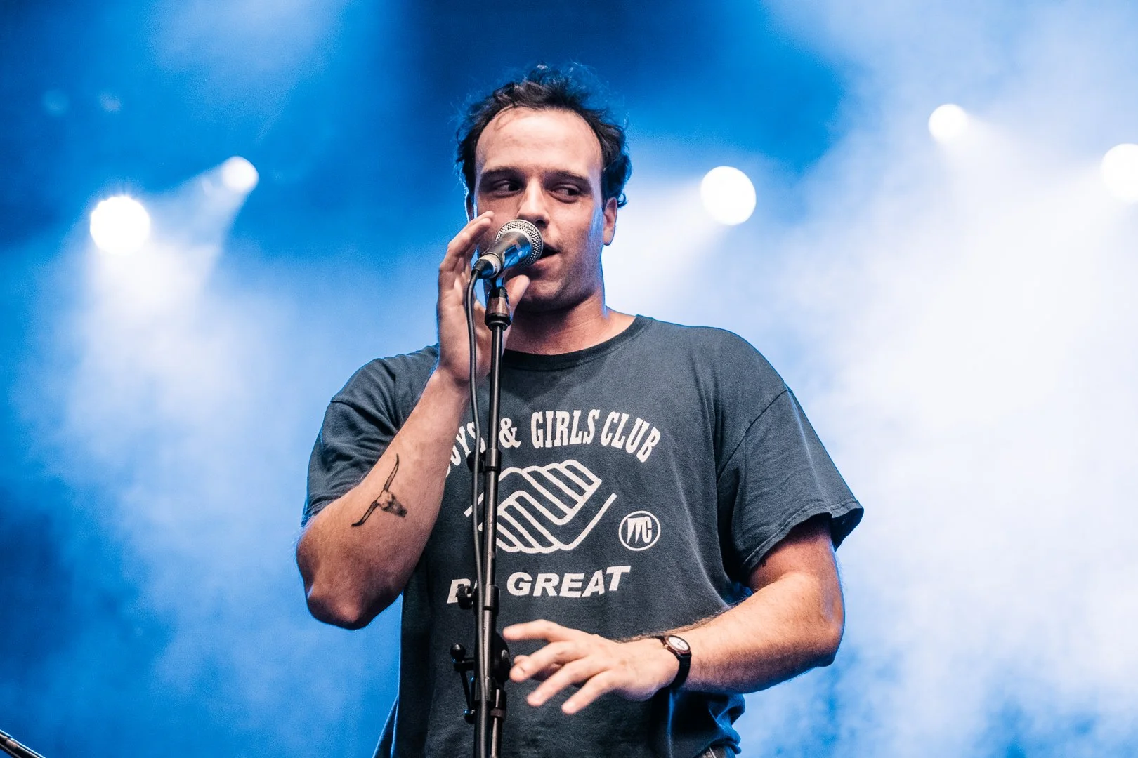 A man with dark hair and a black t-shirt is holding a microphone and singing on stage, with blue stage lighting in the background.