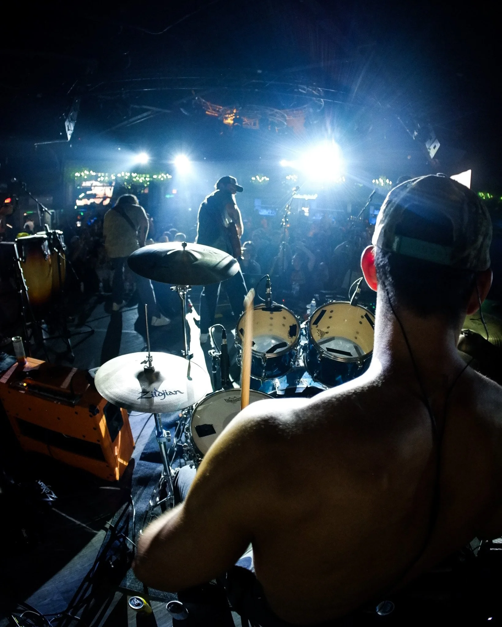 A live music performance at a concert, featuring a drummer in the foreground with a drum set, and silhouettes of band members with guitars and microphones on stage under bright stage lights.