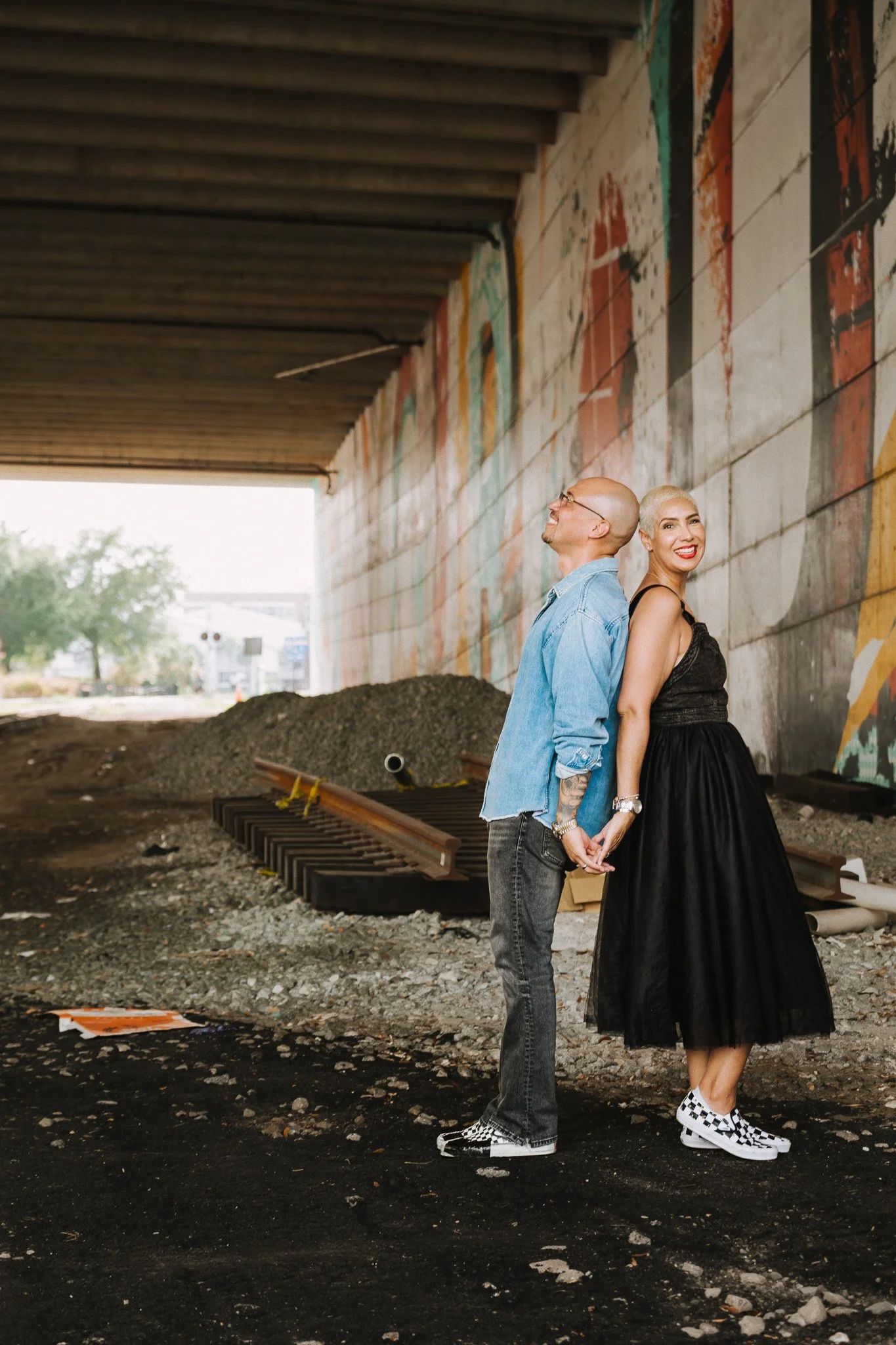 Two people standing back to back under a bridge with colorful murals, holding hands, with construction materials and gravel on the ground.