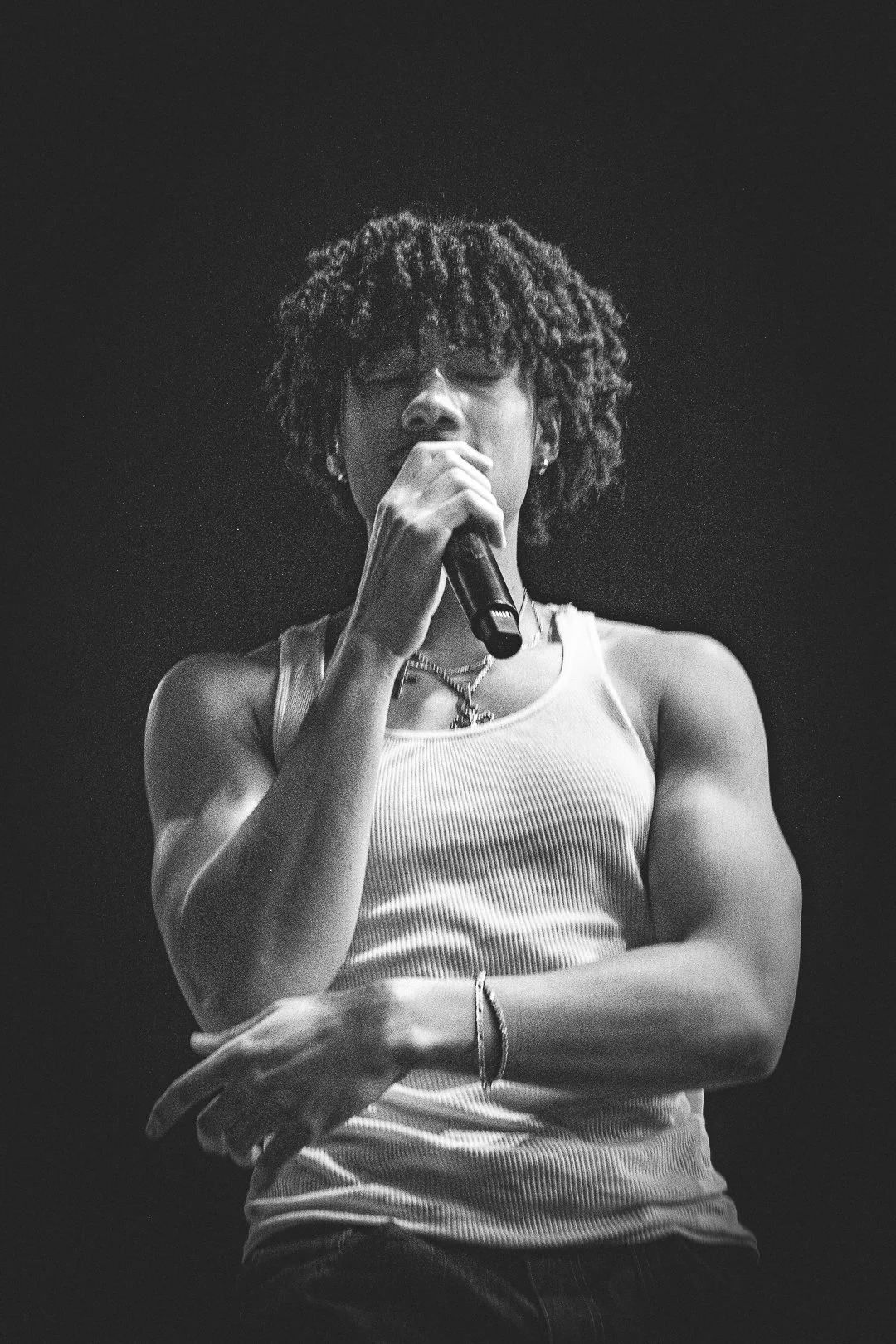 A black-and-white photo of a person singing into a microphone, wearing a tank top and jewelry, with curly hair.