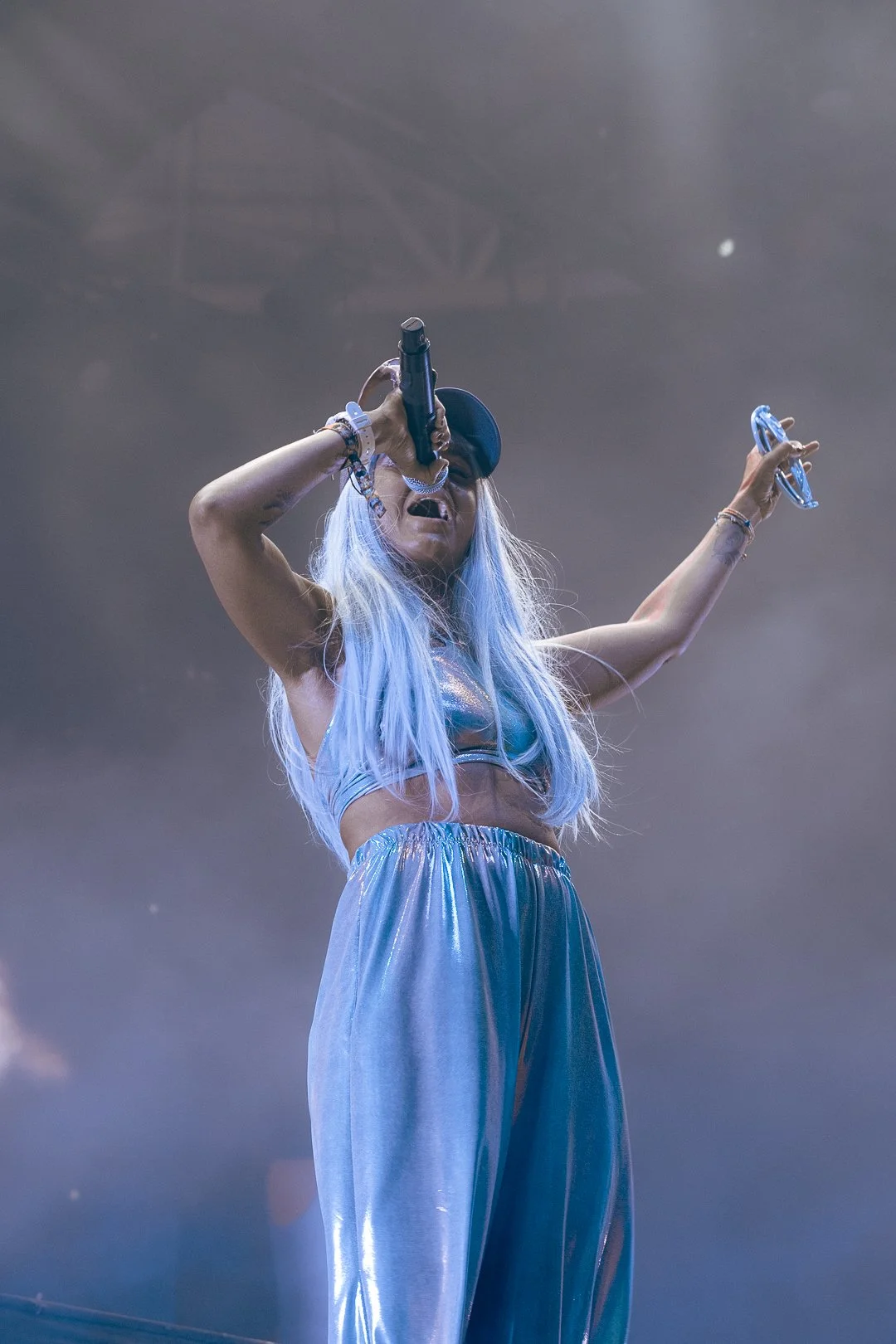 A female performer with long, light blue hair wearing a shiny, silver crop top and matching wide-leg pants, singing into a microphone on stage with a smoky background.