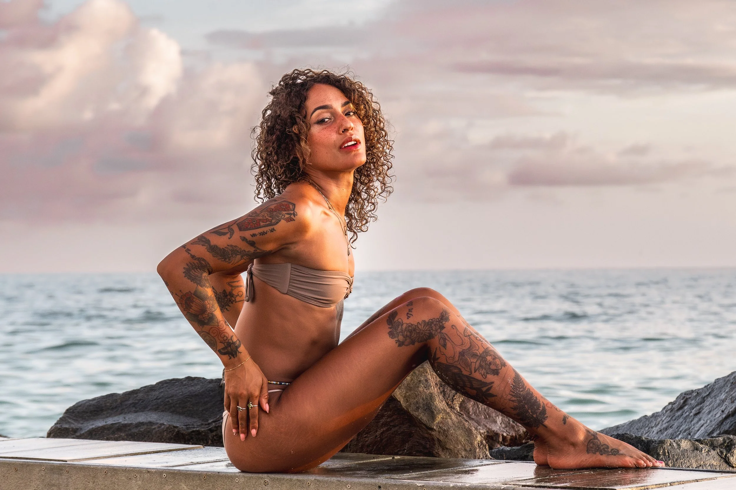 A woman sitting on rocks by the ocean at sunset, wearing a swimsuit and covered in tattoos.