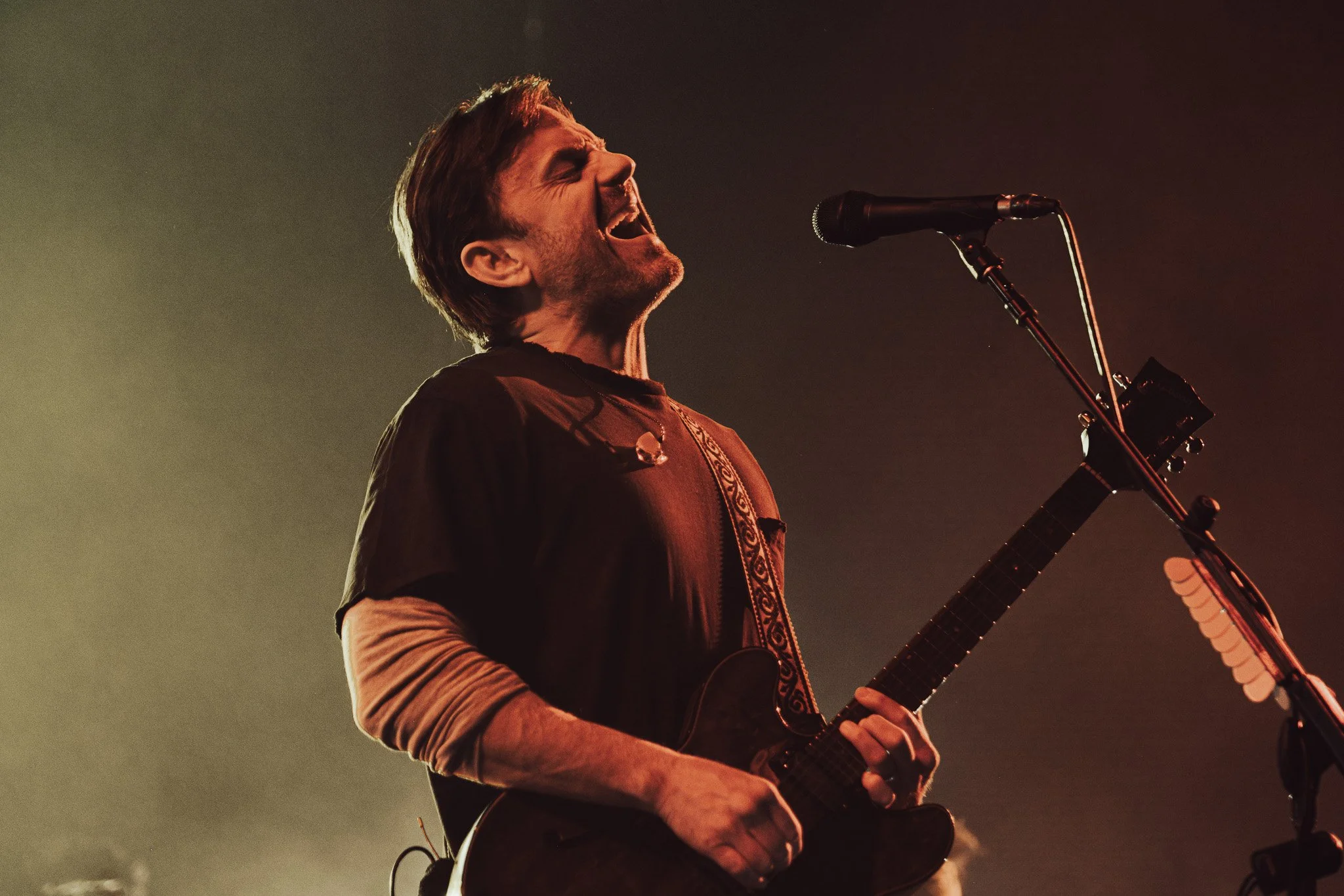 A man with long hair and a beard singing passionately into a microphone while playing an acoustic guitar on stage, illuminated by stage lighting.