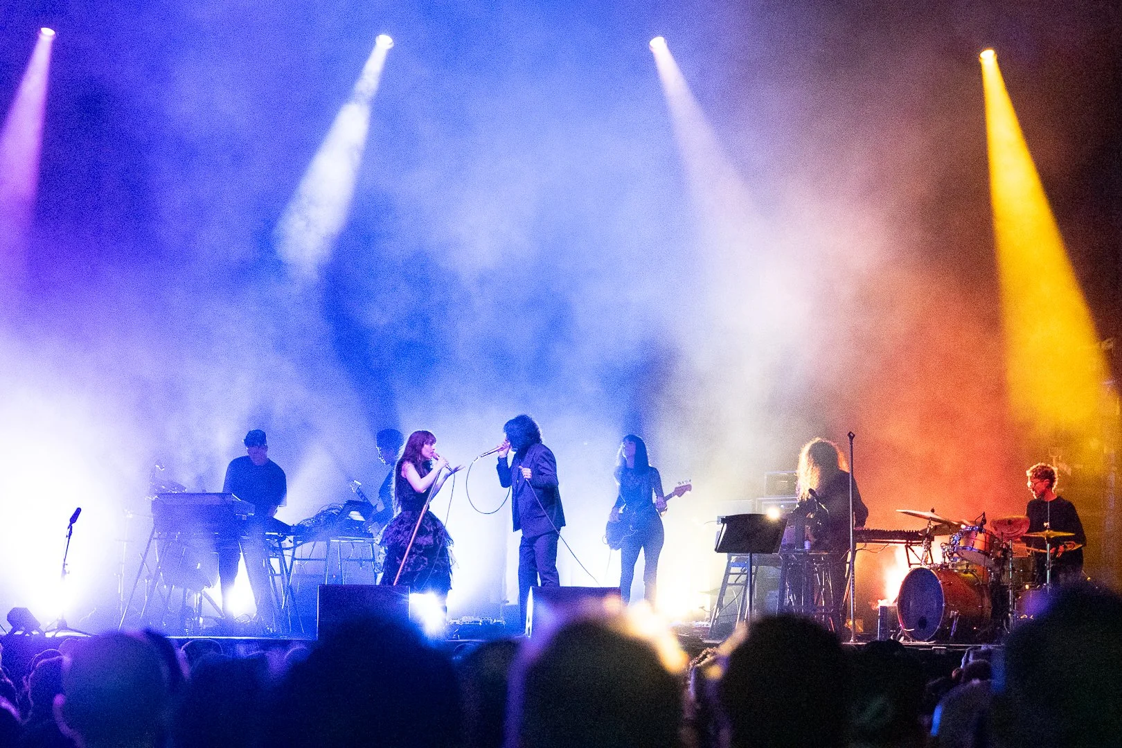 A band performing on stage with colorful lighting and fog effects, featuring singers, guitarists, keyboard players, and a drummer.