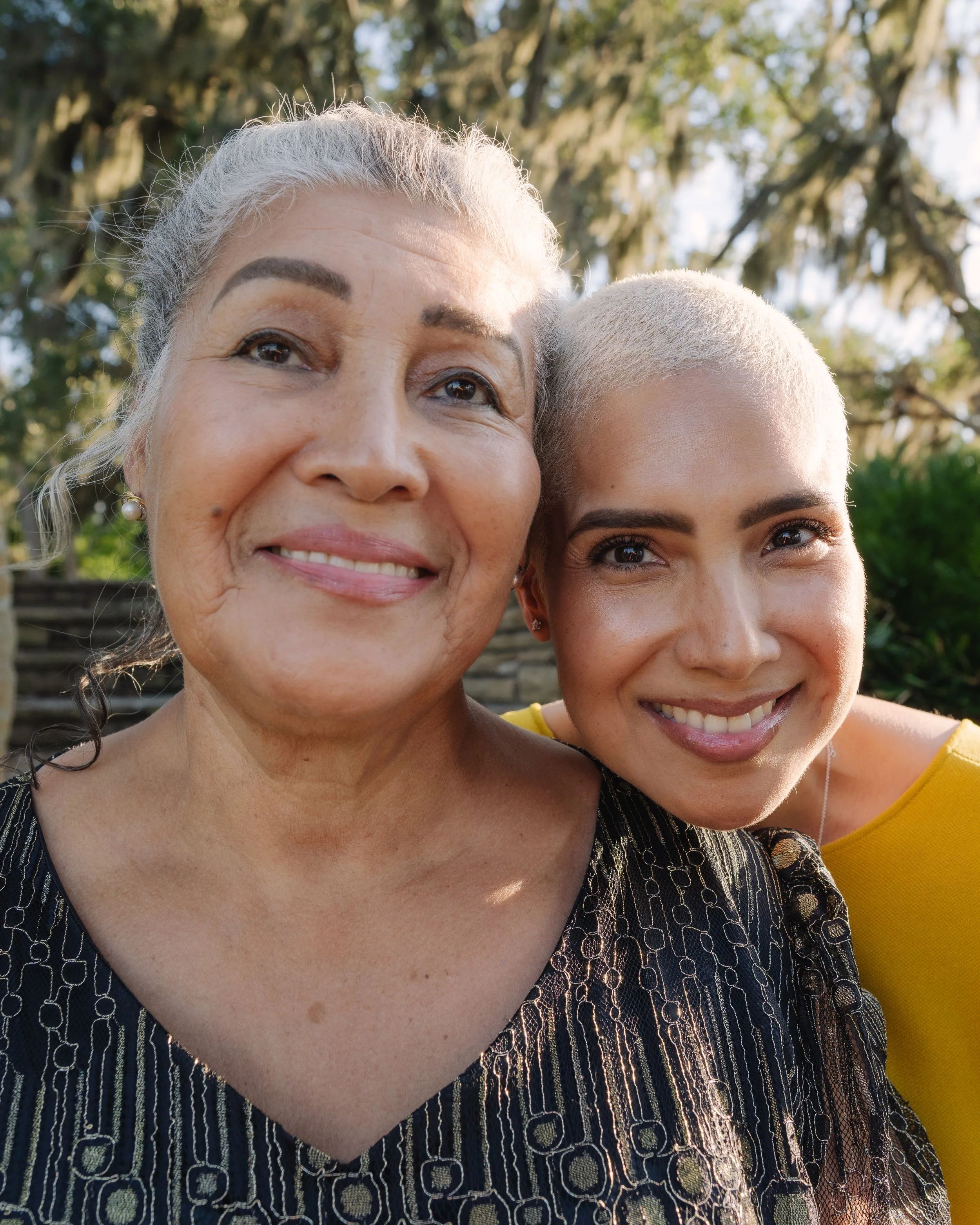 Close-up of two women smiling outdoors with trees in the background. One has gray hair and the other has blonde hair, both with makeup.