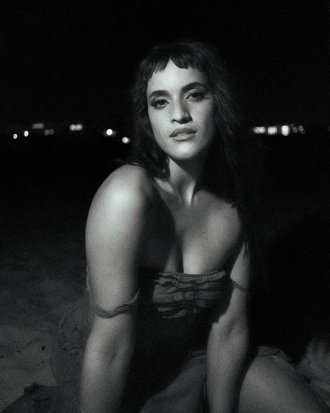 A black and white photo of a woman with dark hair and a nose ring, sitting outdoors at night with city lights in the background.