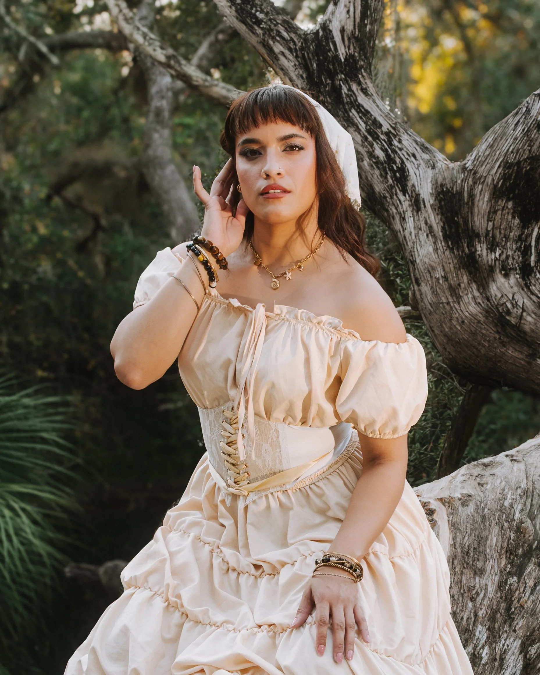 A woman in a vintage-style beige off-the-shoulder dress with puffed sleeves, sitting outdoors near a twisted tree with blurred green foliage and yellow sunlight in the background. She has short brown hair with bangs, jewelry including necklaces and b
