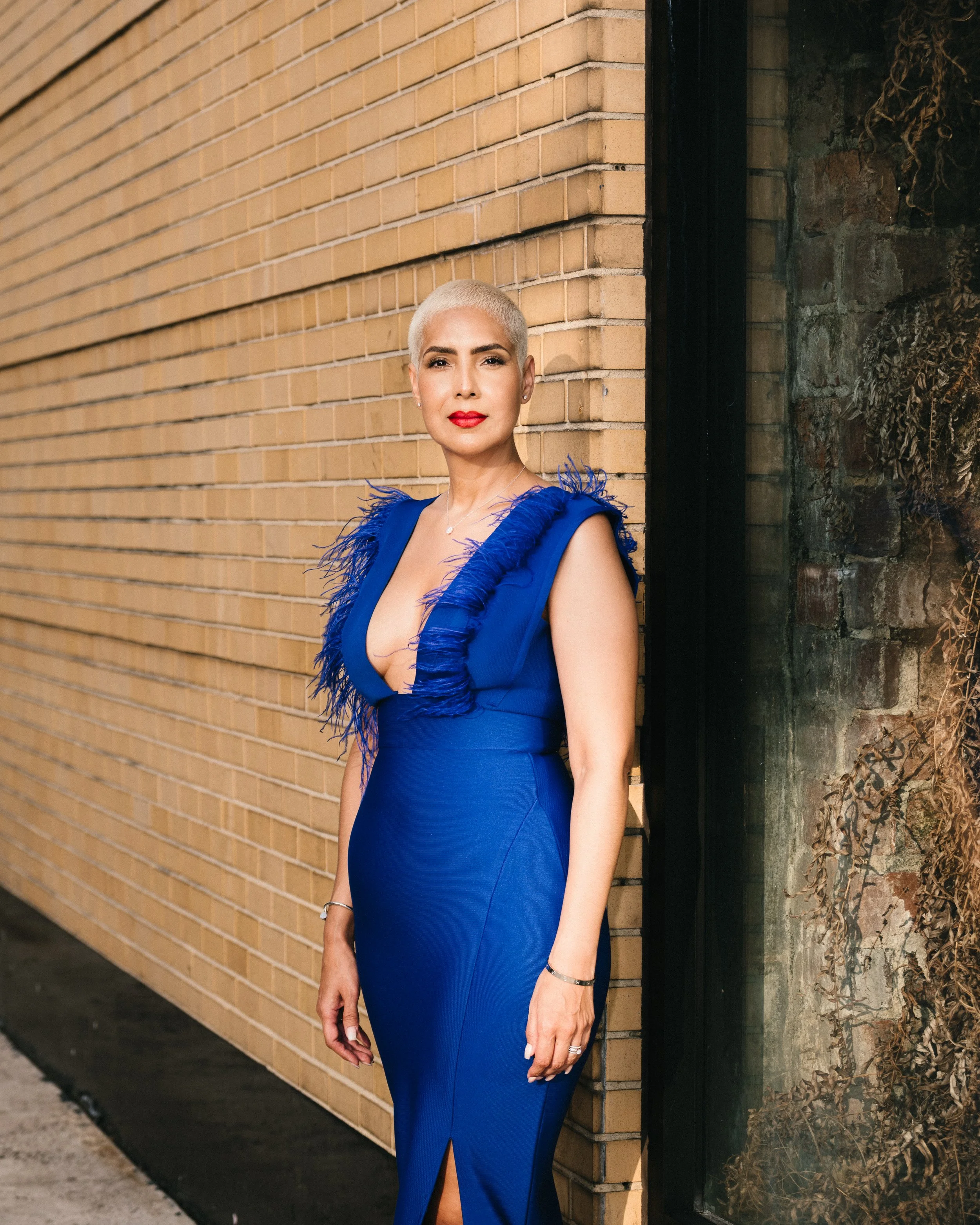 A woman with very short platinum blonde hair and red lipstick standing outdoors against a brick wall. She is wearing a royal blue dress with feathered details on the shoulders and a slit at the front, accessorized with jewelry.
