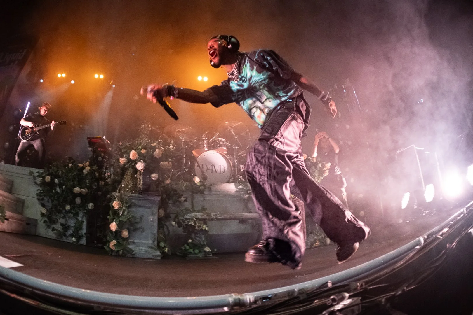 Performer in colorful clothing and jewelry singing on stage during a concert, with musicians playing guitars in the background and stage decorated with flowers and lights.