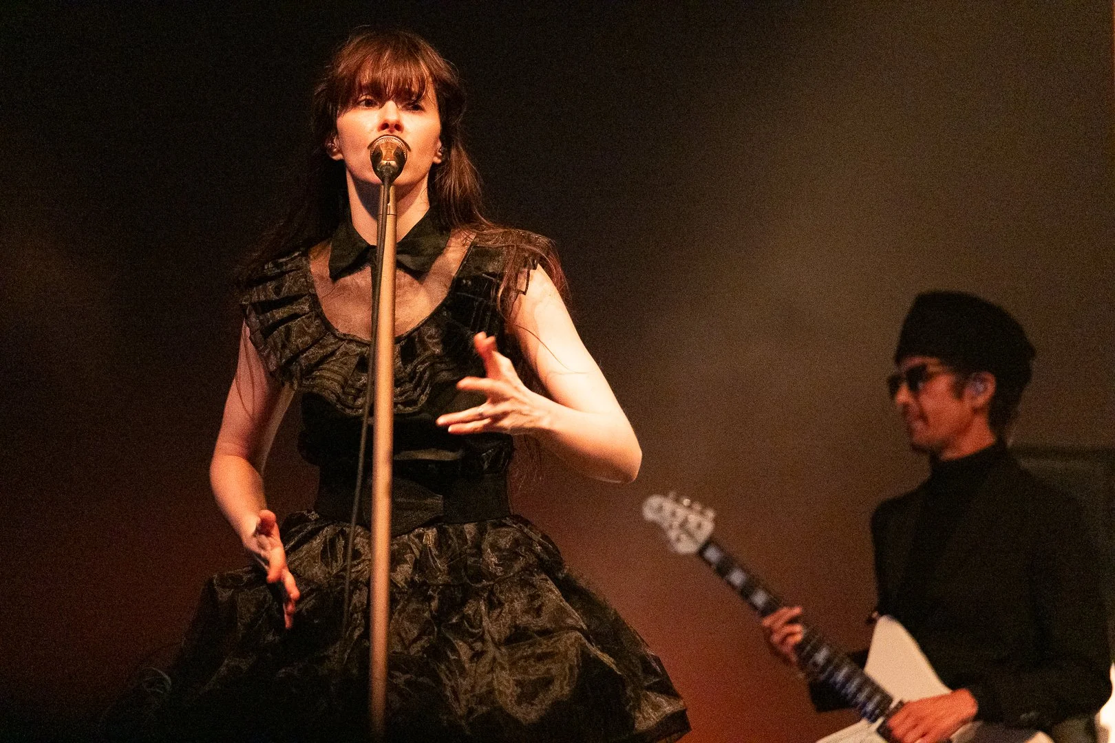 A woman singing into a microphone on stage, wearing an elaborate black dress with ruffles, and a man in the background playing an electric guitar, wearing glasses and a black outfit.