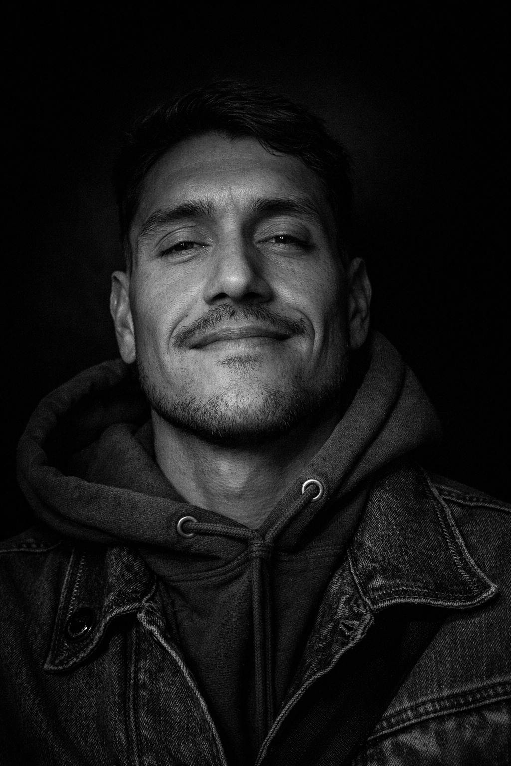 Black and white portrait of a smiling man wearing a hoodie and denim jacket.