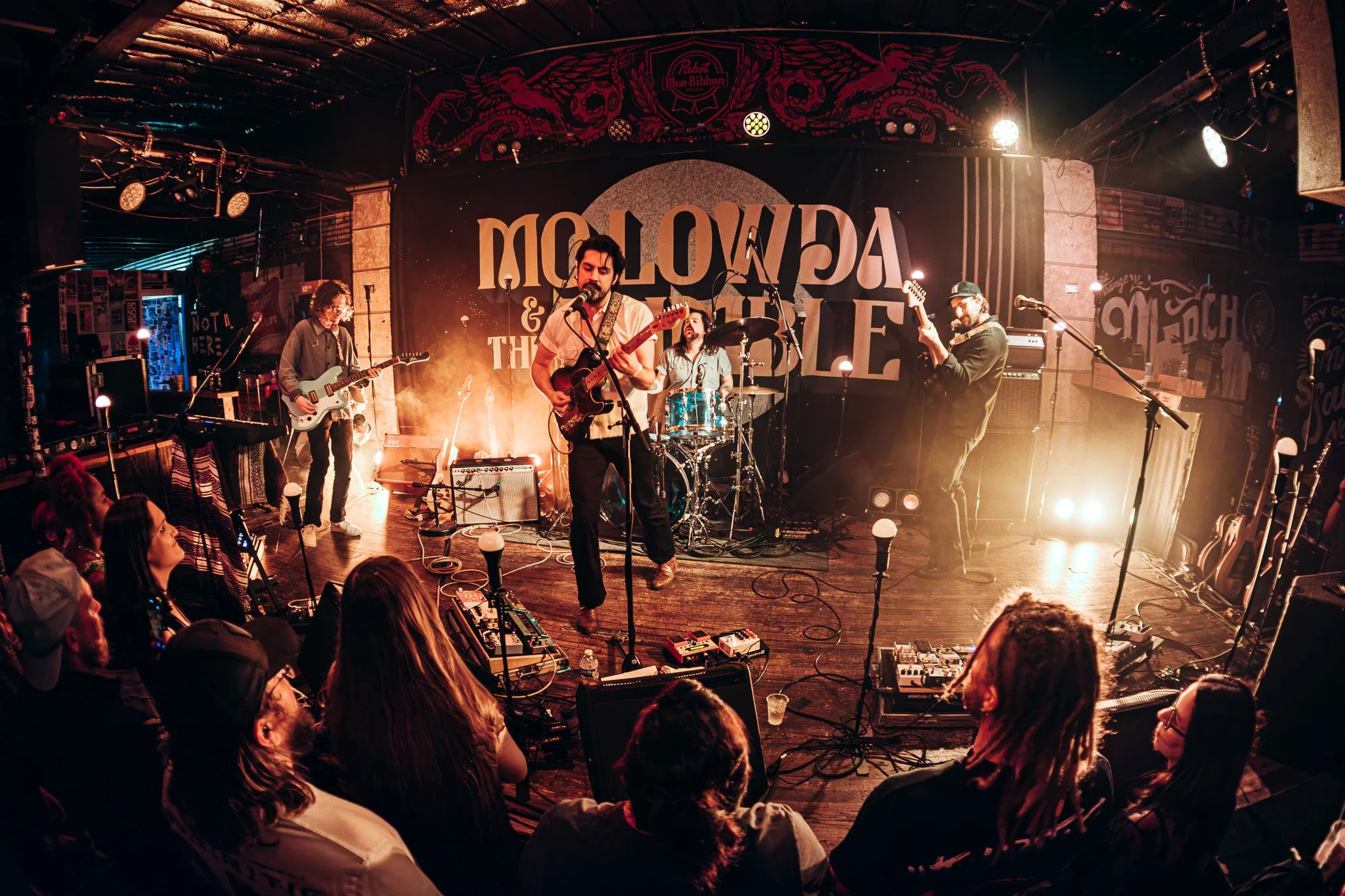 A band performs live on stage in a dimly lit venue. The backdrop displays the words 'DIOLOWA & THE...'. The audience is seated and watching the performance, with some visible in the foreground.