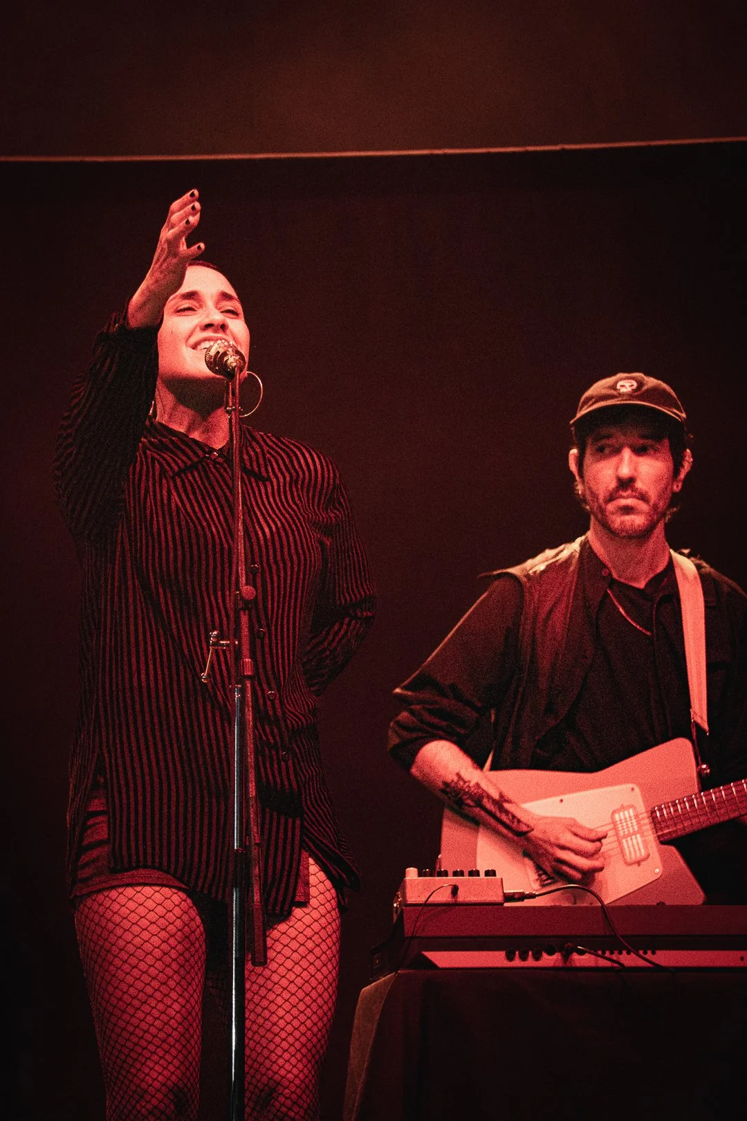 A woman with dark hair singing into a microphone, wearing a striped shirt and fishnet stockings, and a man with dark hair wearing a cap and playing an electric guitar or keyboard, on a dark stage with red lighting.