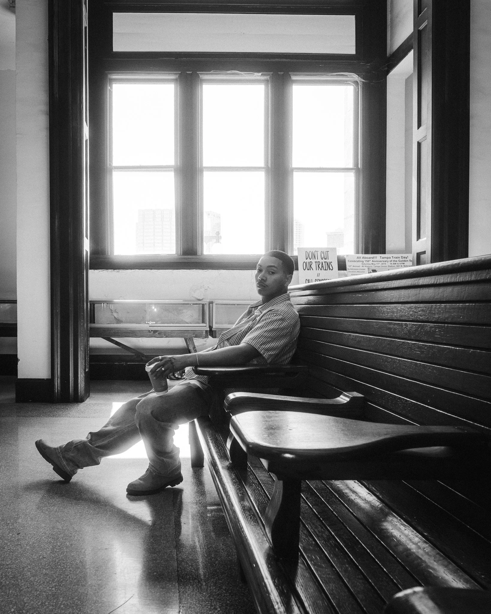 A young man sitting on a wooden bench inside a room with large windows, holding a drink container, with signs in the background protesting against cuts to transit services.