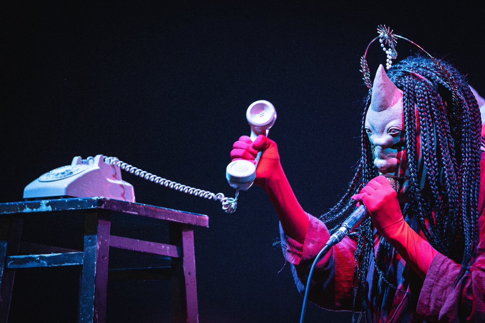 A person dressed in a cat-like costume with a detailed mask, long braided hair, and wearing red gloves, is holding a vintage rotary phone to their ear and speaking into a microphone. The scene is dark with stage lighting.
