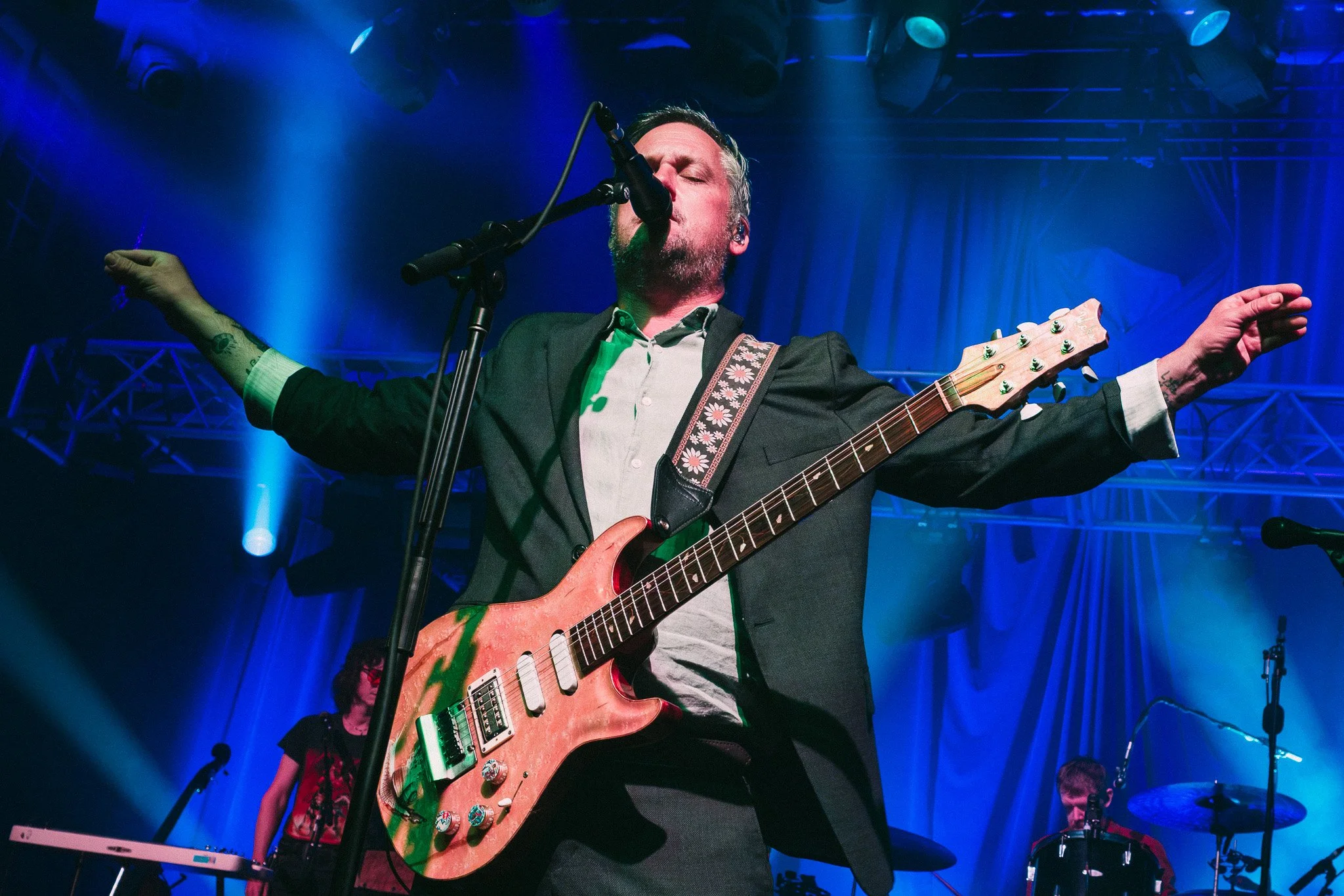 A male musician with tattoos on his arms, wearing a dark blazer and light-colored shirt, performing on stage with a pink electric guitar and singing into a microphone. Blue stage lighting and a band member playing keyboard can be seen in the backgrou