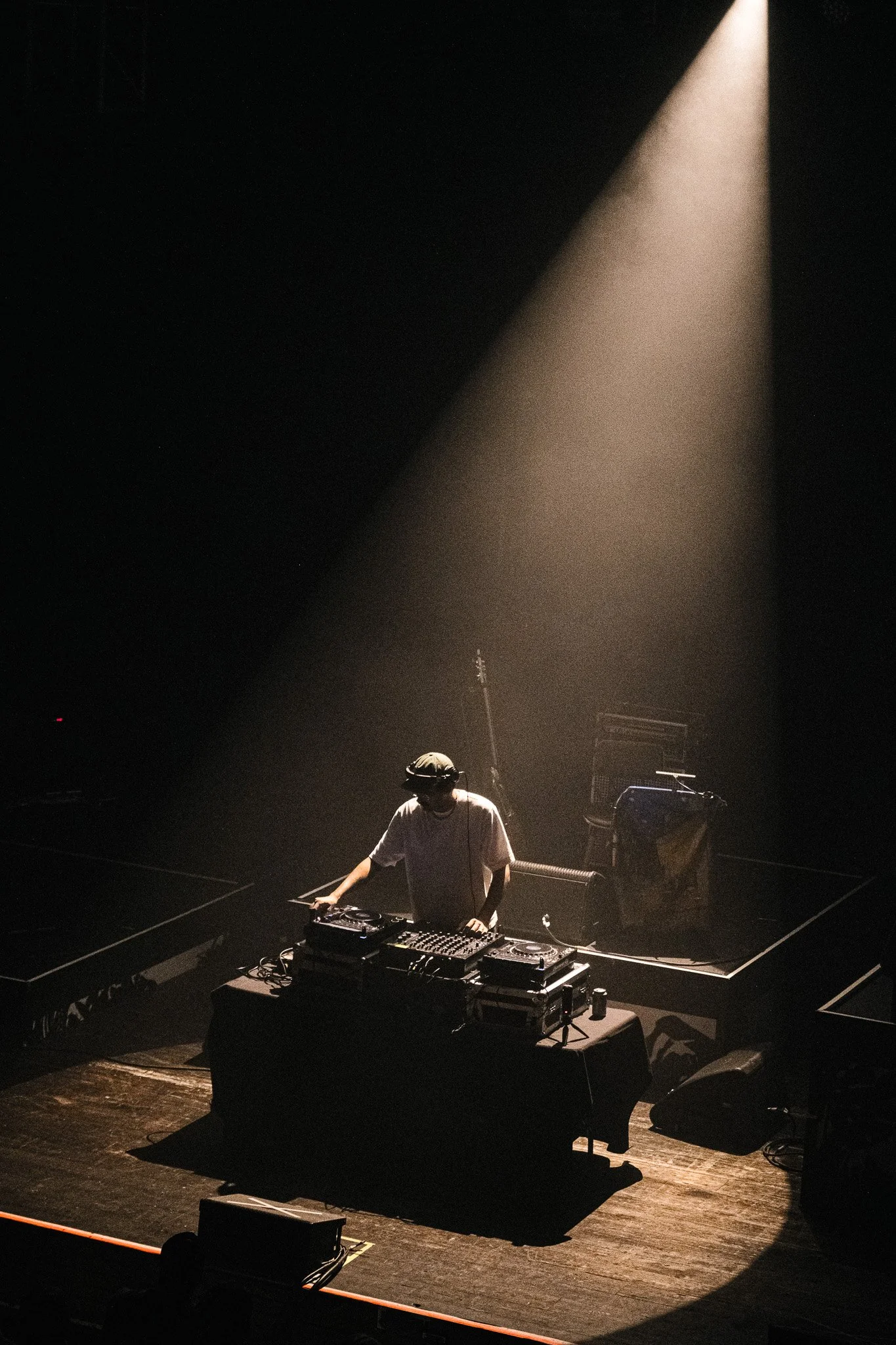A DJ wearing a white shirt and headphones is performing behind a DJ booth on a dark stage, illuminated by a single spotlight.