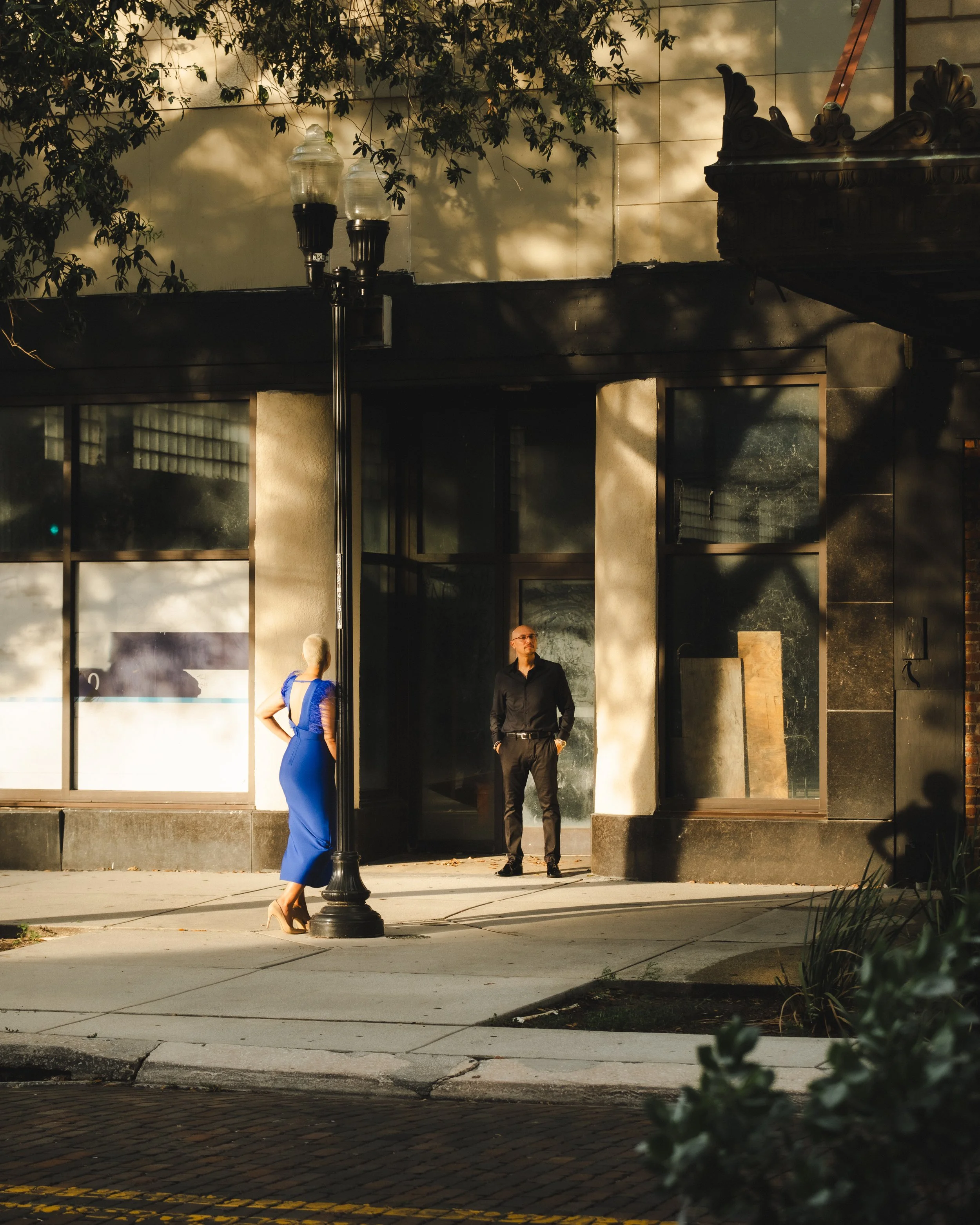 Two people stand outside a building with large glass windows, one woman in a blue dress with high heels and a man in black, during late afternoon or early evening with shadows cast on the building.
