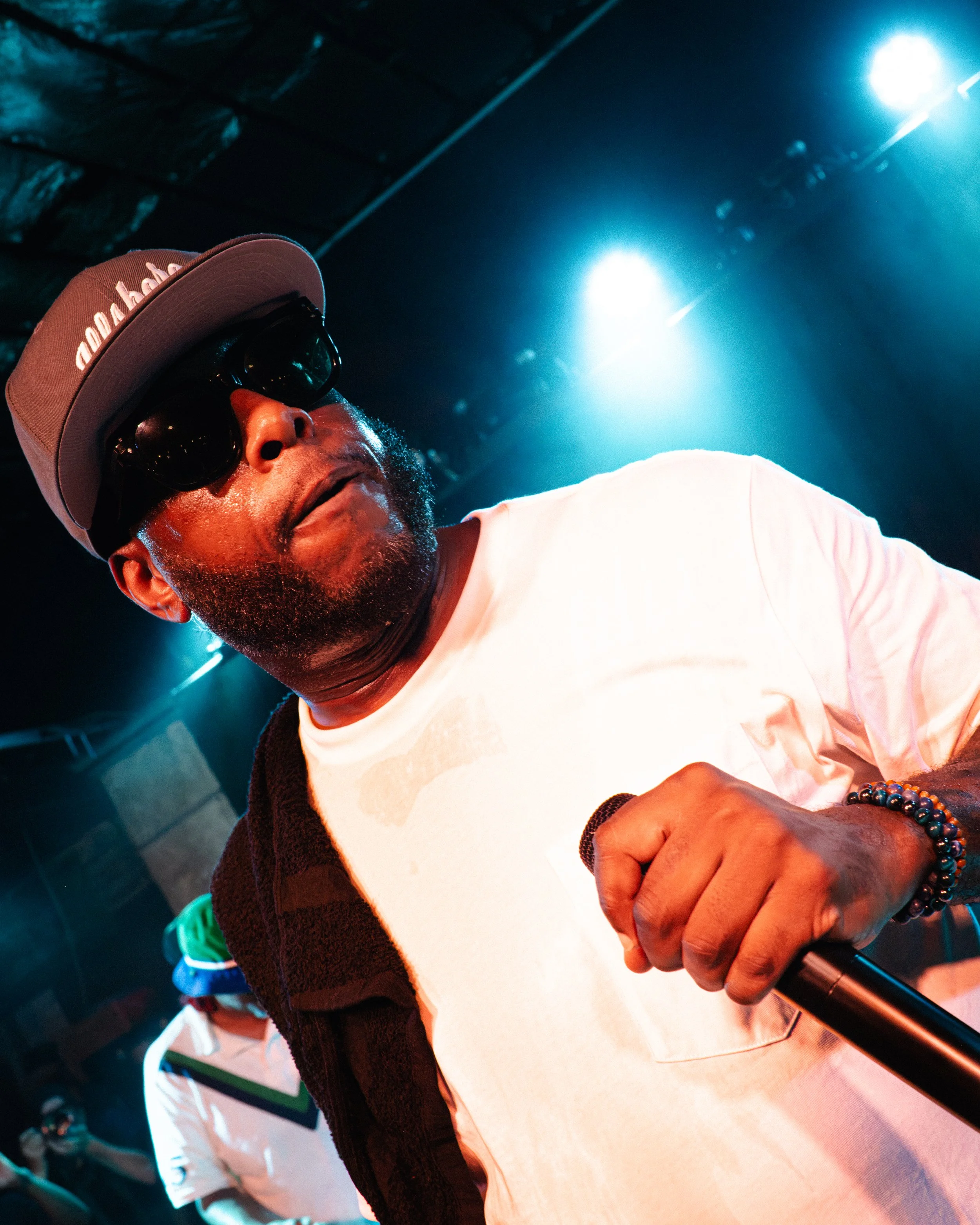 A close-up of a man wearing a light pink baseball cap with white text, black sunglasses, and a white T-shirt, holding a microphone. The background is illuminated with blue stage lights.