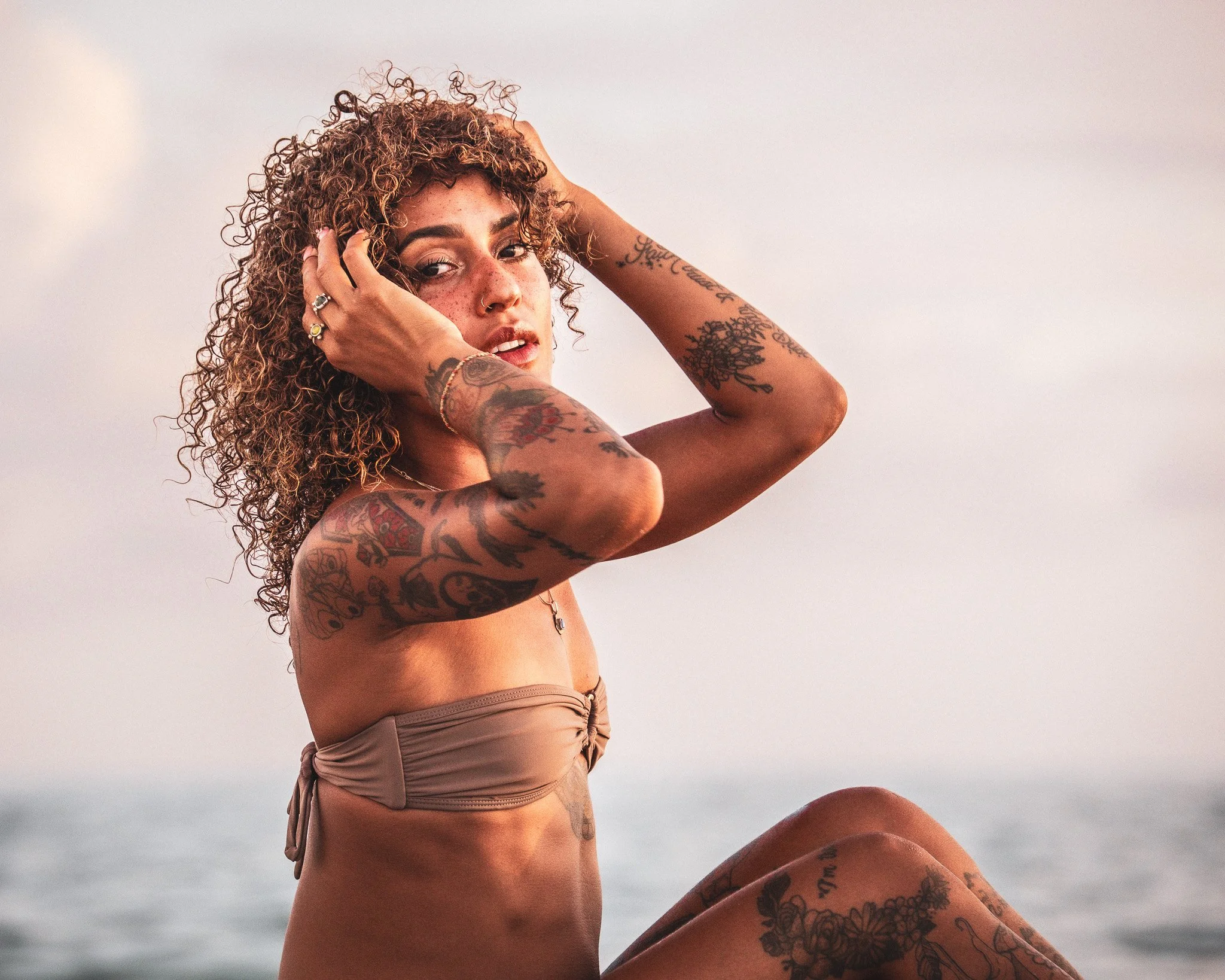 A woman with curly hair and tattoos on her arms and body, wearing beige shorts, sitting outdoors during sunset.