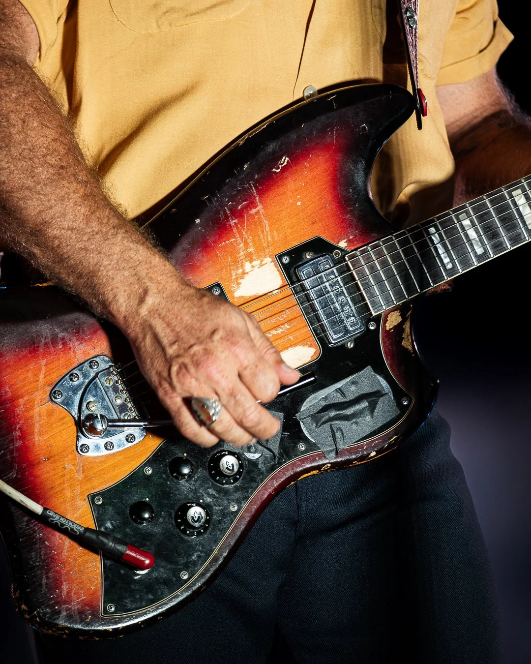 Person playing a vintage, worn electric guitar with a sunburst finish, black pickguard, and multiple control knobs.