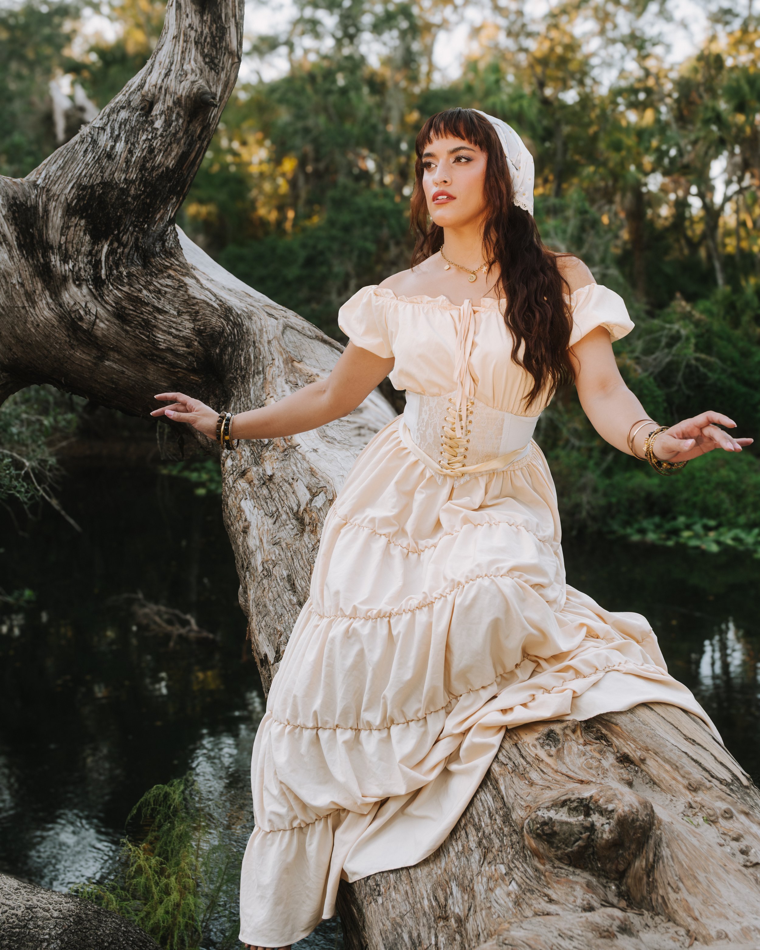 A woman in a vintage cream-colored dress and headscarf sits on a fallen tree trunk near a river in a forest with autumn-colored trees.