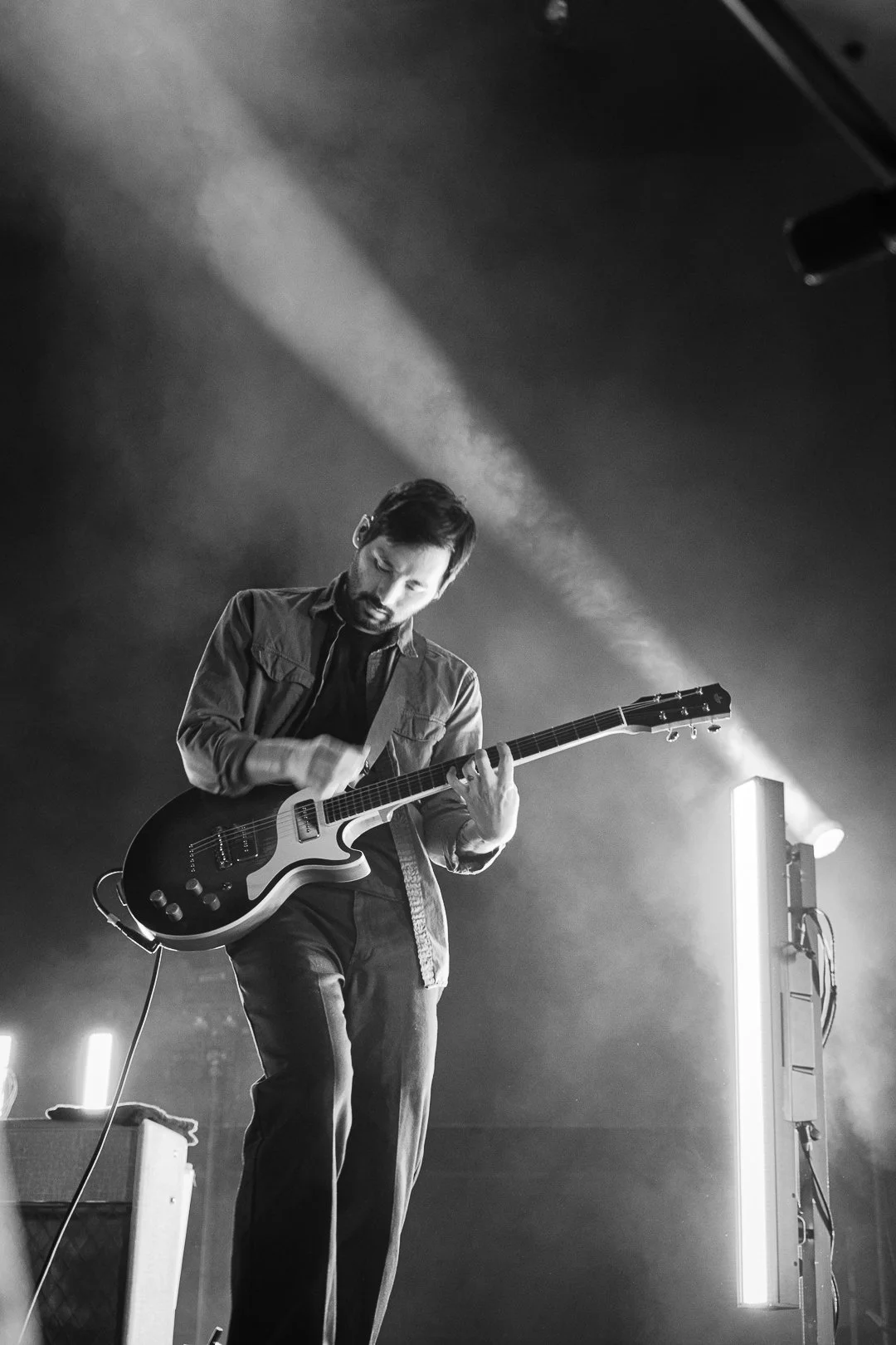 Black and white photo of a man playing an electric guitar on stage, illuminated by stage lights, with fog or smoke in the background.