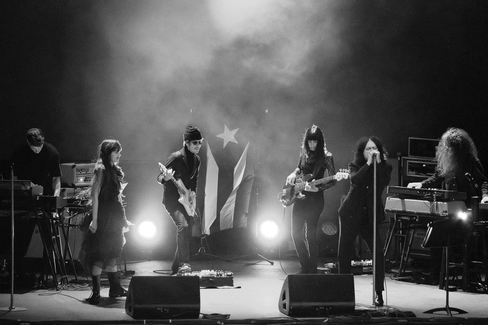 A black-and-white photo of a band performing on stage with five members. Two women are playing keyboards on the left and right, a woman is singing into a microphone in the center, a man is playing an electric guitar, and another woman is playing an e