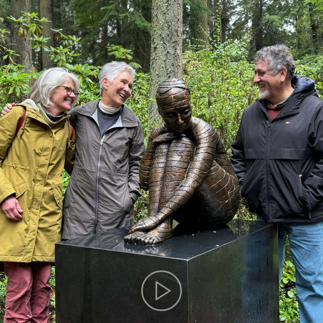Three women and one man smiling and standing around a bronze sculpture of a girl with her knees drawn up to her chest, in a forest setting.