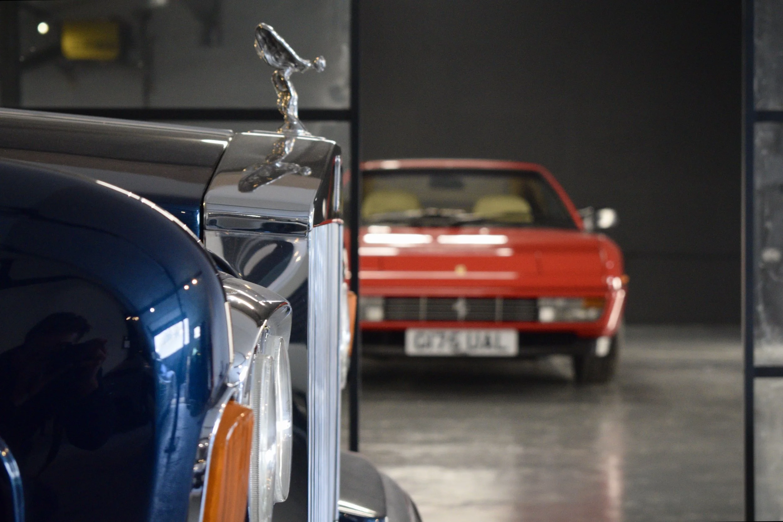 Close-up of classic cars parked indoors, with a red car visible in the background and a metallic hood ornament in the foreground.