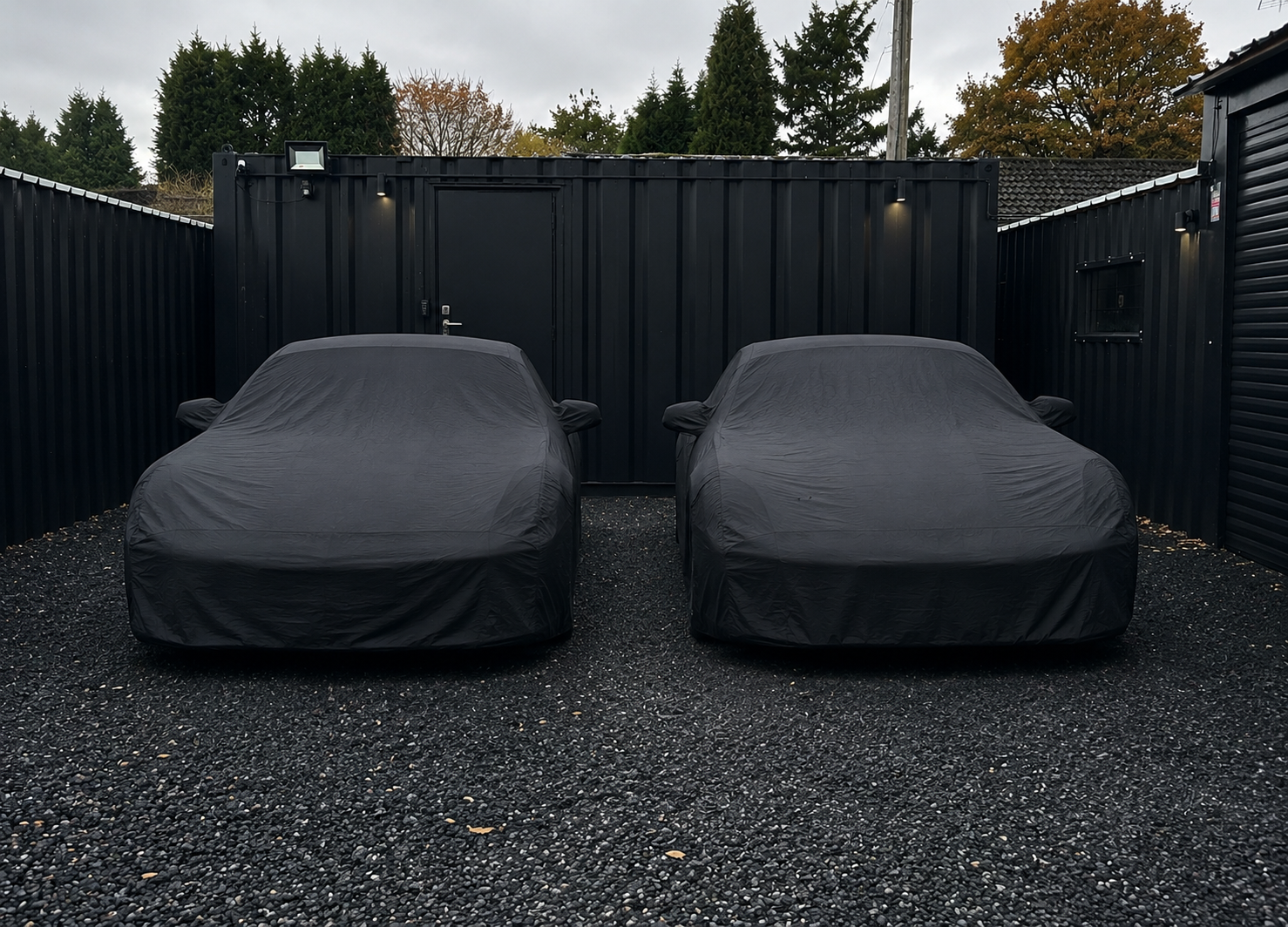 Two cars covered with black car covers parked side by side on a gravel surface in front of a black metal fence with trees in the background.