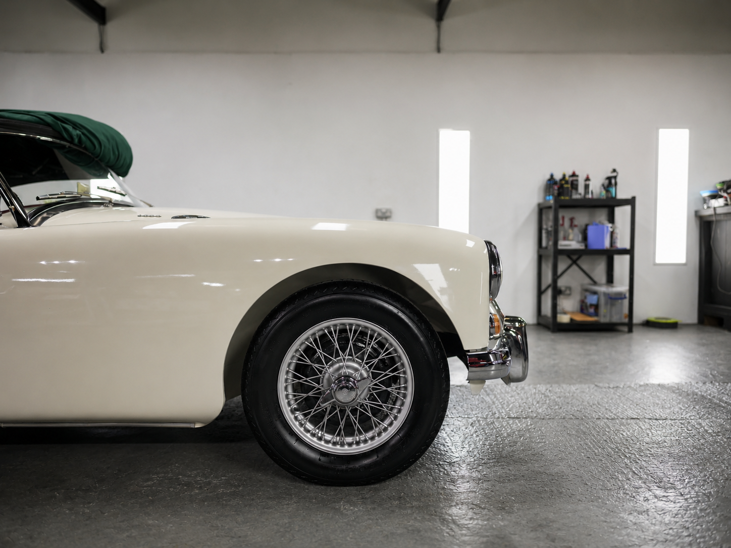 Part of a vintage cream-colored car with wire-spoke wheel in a garage, background includes shelves with tools and supplies.