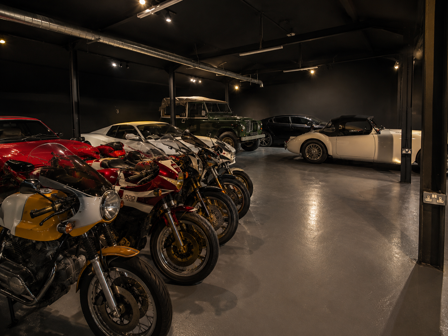 A collection of vintage motorcycles and classic cars in a dimly lit garage or showroom. The motorcycles are lined up in the foreground, with several sports cars and a green utility vehicle in the background.