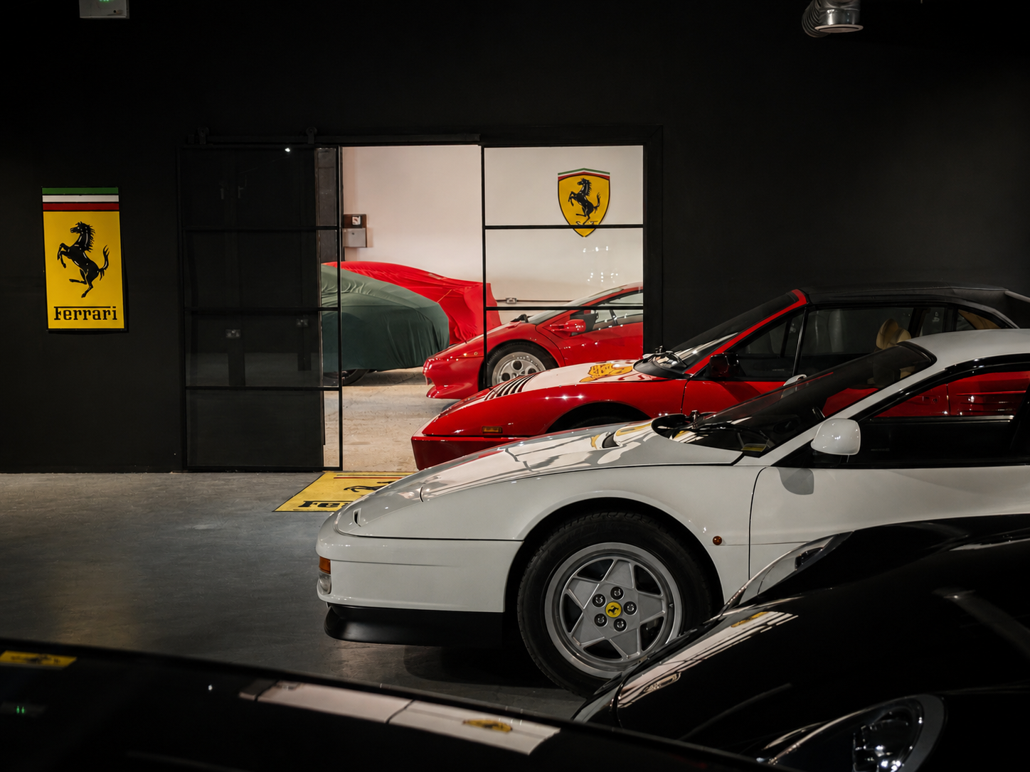 A garage with multiple Ferrari cars, including a visible white Ferrari in the front, a red Ferrari behind it, and another red Ferrari in a glass-walled room, with Ferrari and shield logos on the black walls.