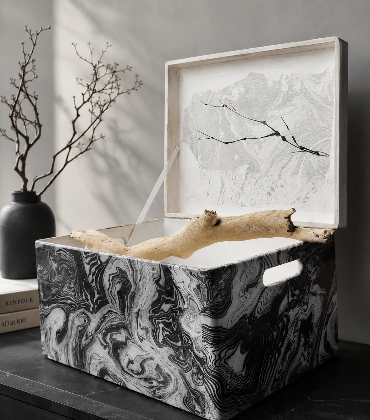 Decorative box with black and white marble swirl pattern, open to reveal a piece of driftwood inside, with a leafy branch in a black vase and a book in the background.