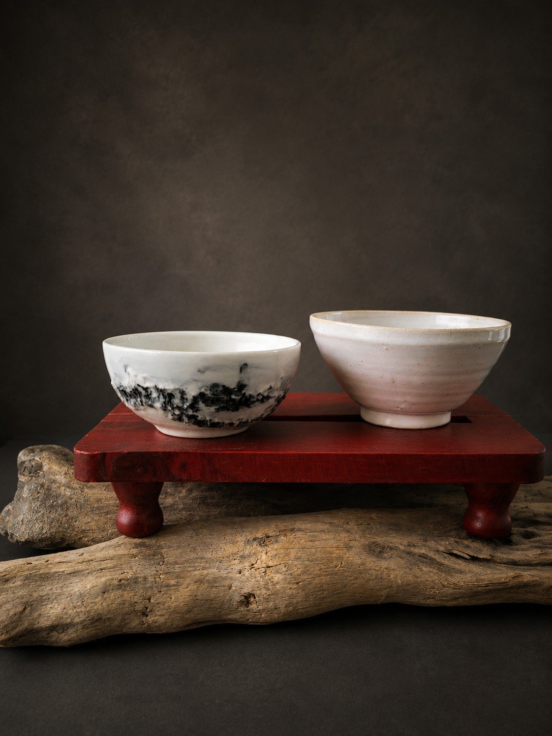 Two ceramic bowls on a red wooden tray, resting on a piece of driftwood. The bowl on the left is white with black marbling, and the one on the right is plain white with subtle horizontal lines.