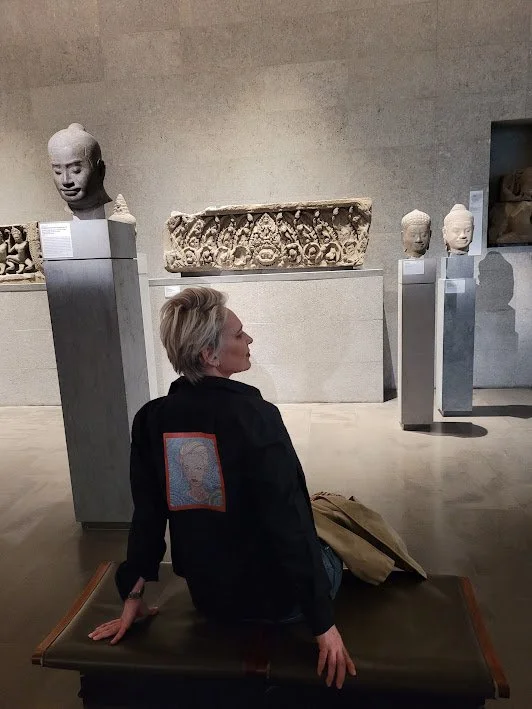 A woman with short blonde hair is seated on a bench in an art museum, surrounded by ancient stone sculptures and busts displayed on pedestals and shelves.