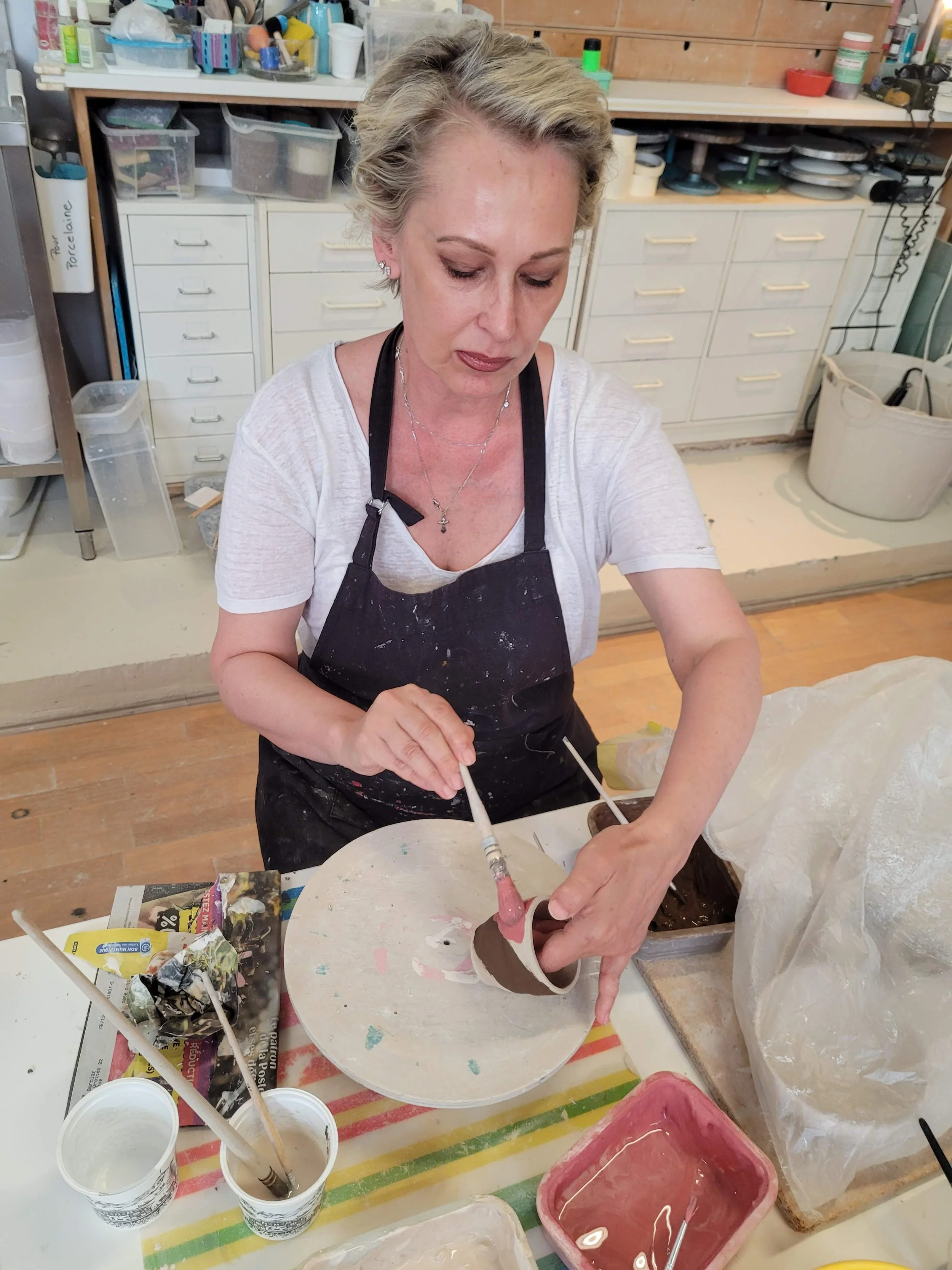 A woman working on a pottery project at a table in a ceramics studio, using a brush and a cylindrical object with clay. The table has various containers of water, paint, and clay. Shelves with art supplies are in the background.