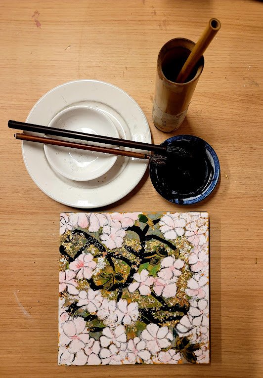 A small square canvas with pink and white cherry blossom flowers and black and gold accents, placed on a wooden table. Nearby are two small dishes, one white and empty, the other dark and containing dark-colored liquid, with a pair of chopsticks resting across the white dish. A tall cylindrical container holds a yellow straw and a brown object.