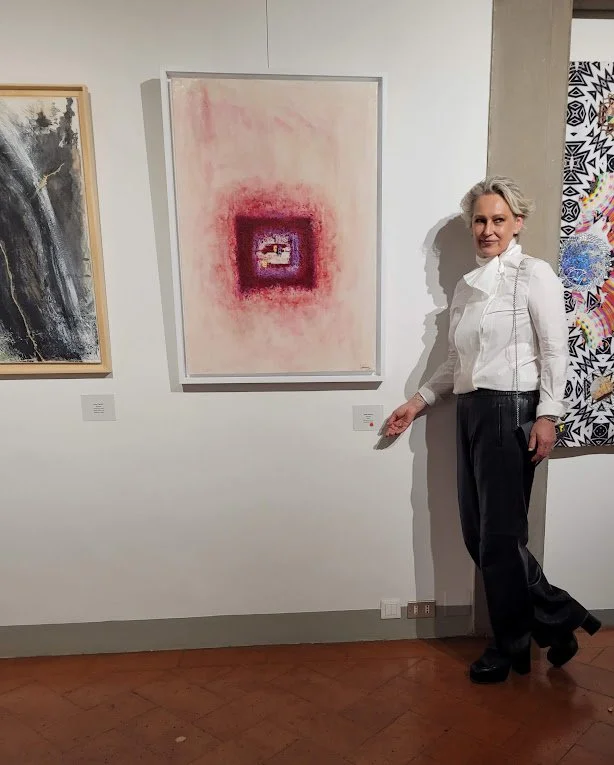 A woman with short gray hair wearing a white blouse and black pants standing in an art gallery next to a large abstract painting with pink, purple, and red hues, pointing to a label beneath the artwork.