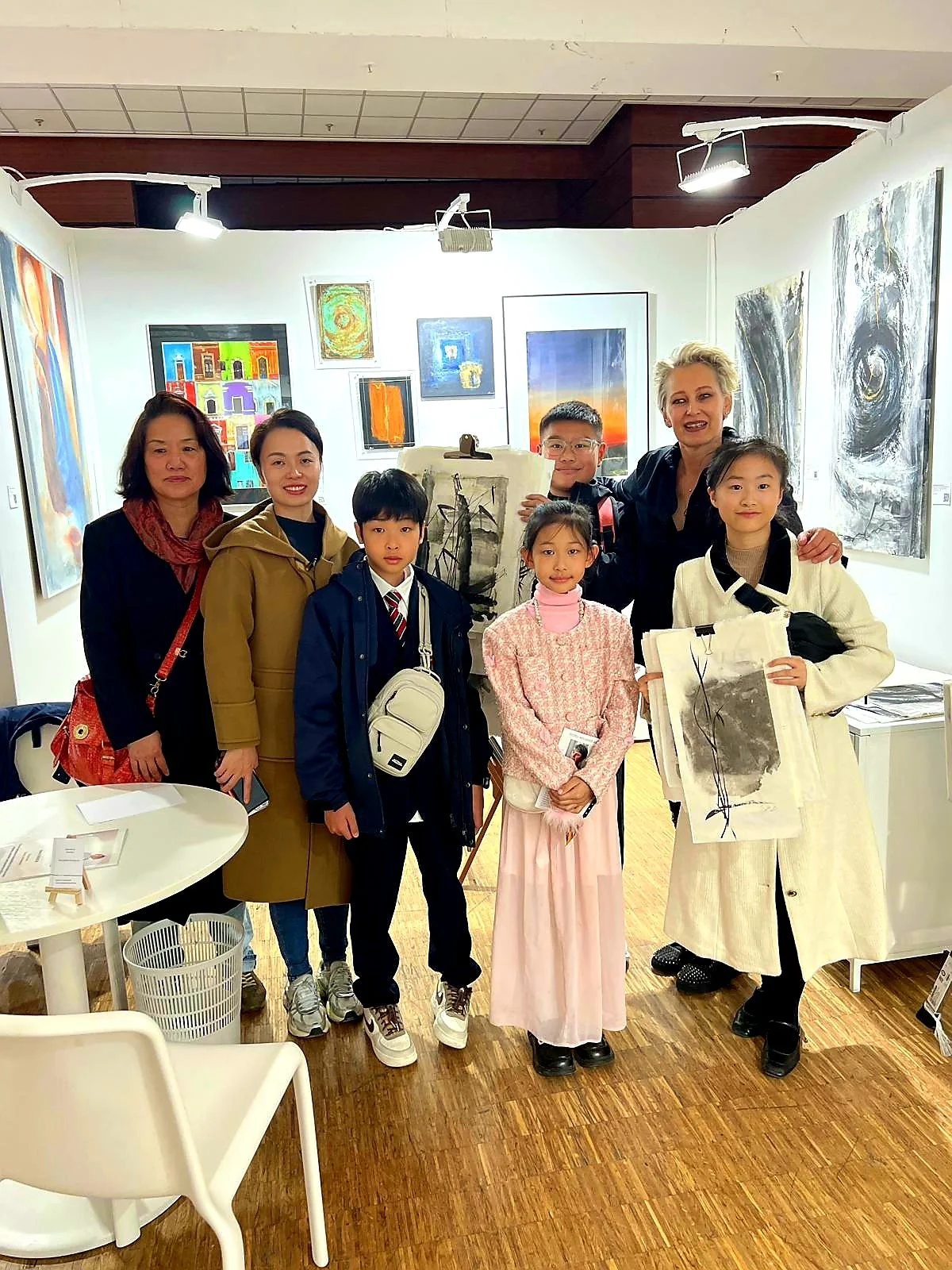 A diverse group of people, including children and adults, gathered in an art gallery with various artworks displayed on the walls. They are posing for a photo, with several smiling and holding art pieces or books.