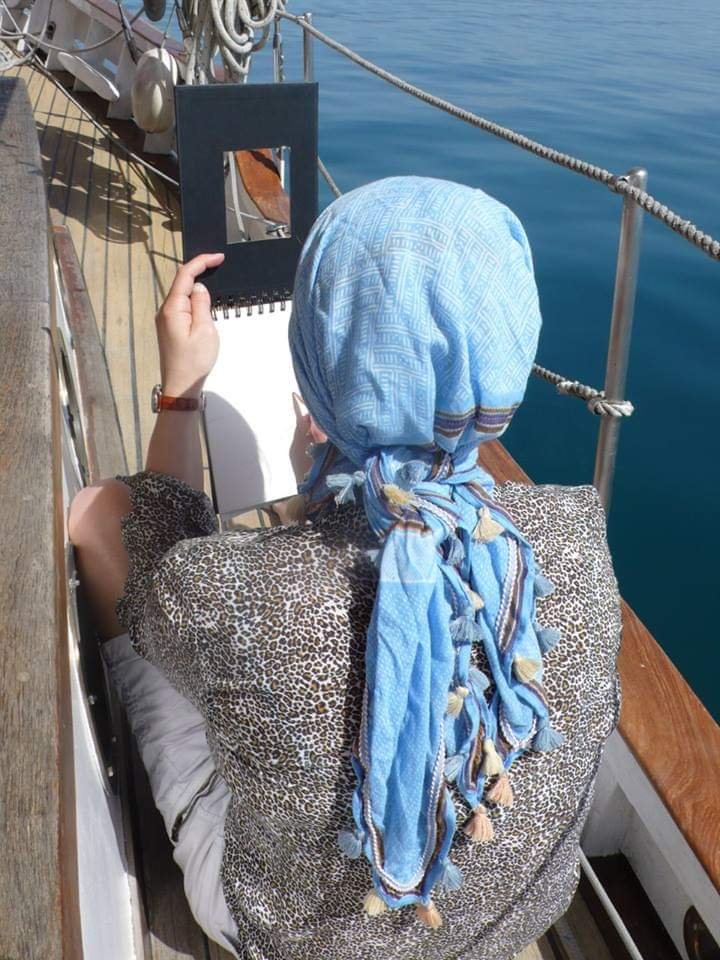 Person sitting on a boat, wearing a blue headscarf, leopard-print top, and light-colored shorts, drawing or writing in a sketchbook with a mirror in hand, overlooking calm blue water.