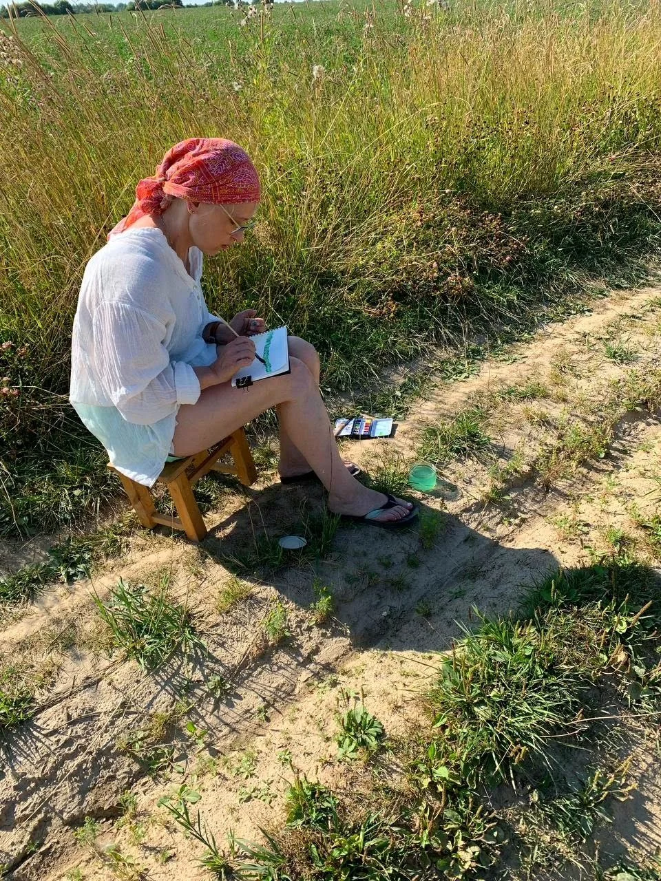 A woman sitting on a small wooden stool in a field of tall grass, drawing or painting on a small canvas with art supplies nearby in the dirt.