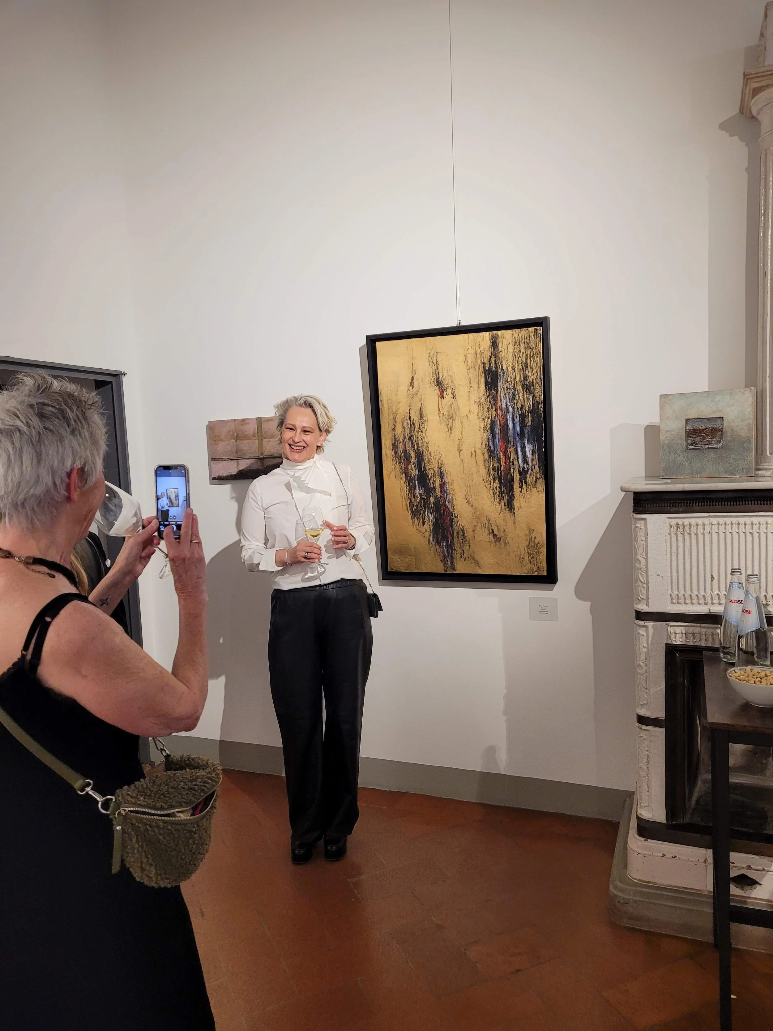 A woman in a white blouse and black pants is smiling while holding a glass of wine, standing in an art gallery. Other people are taking photos of her, and artworks are displayed on the wall behind her.