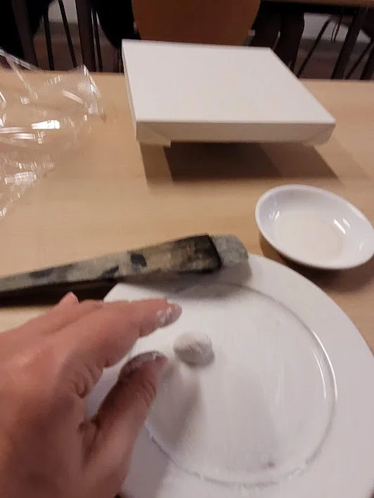 A hand shaping dough on a white plate at a table with a bread knife, a small empty dish, and a container of paper bags in the background.