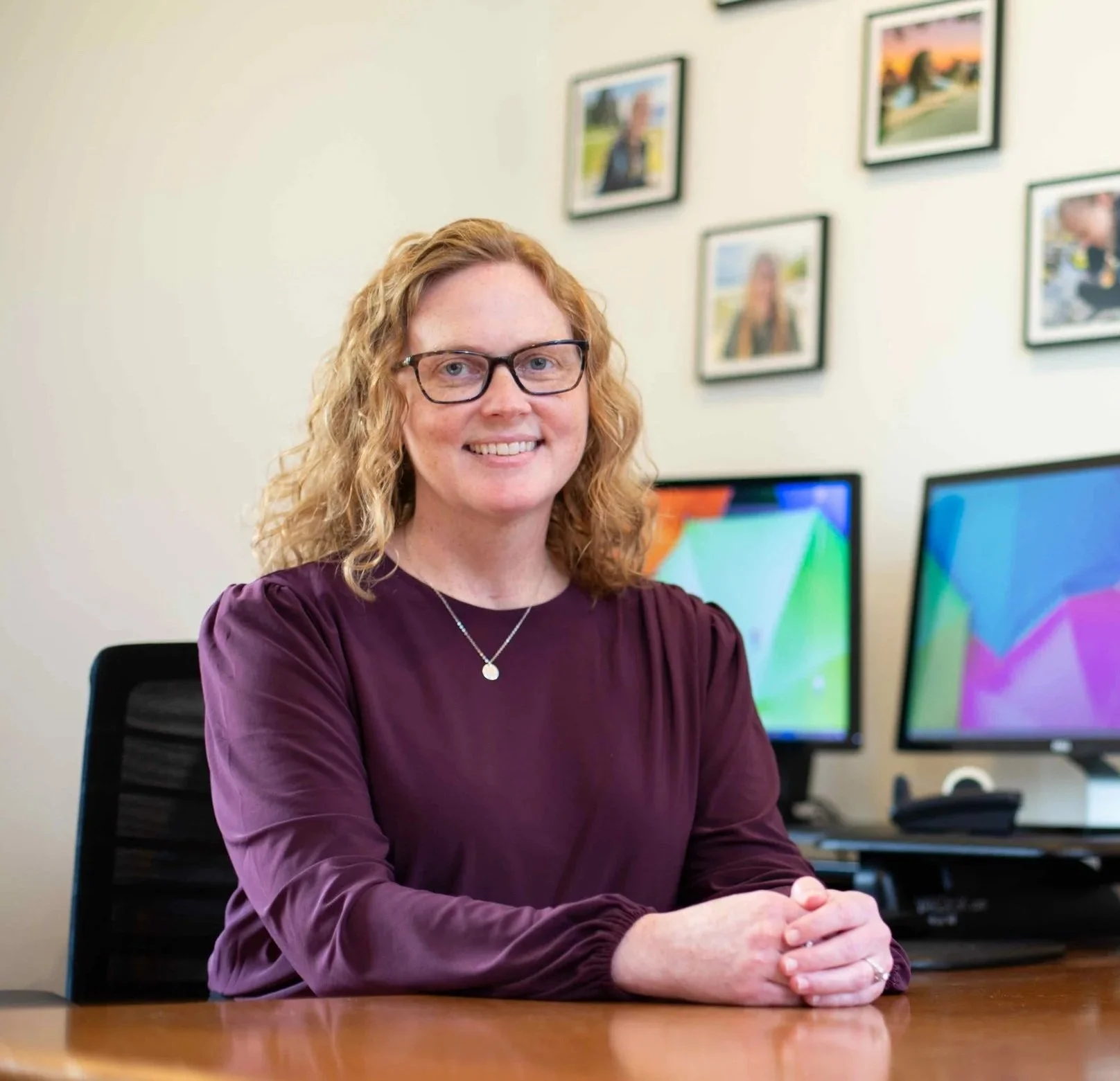 A woman with curly red hair, wearing glasses and a purple long-sleeve top, is sitting at a desk with her hands clasped. Behind her is a wall with framed photos and two computer monitors showing colorful patterns.